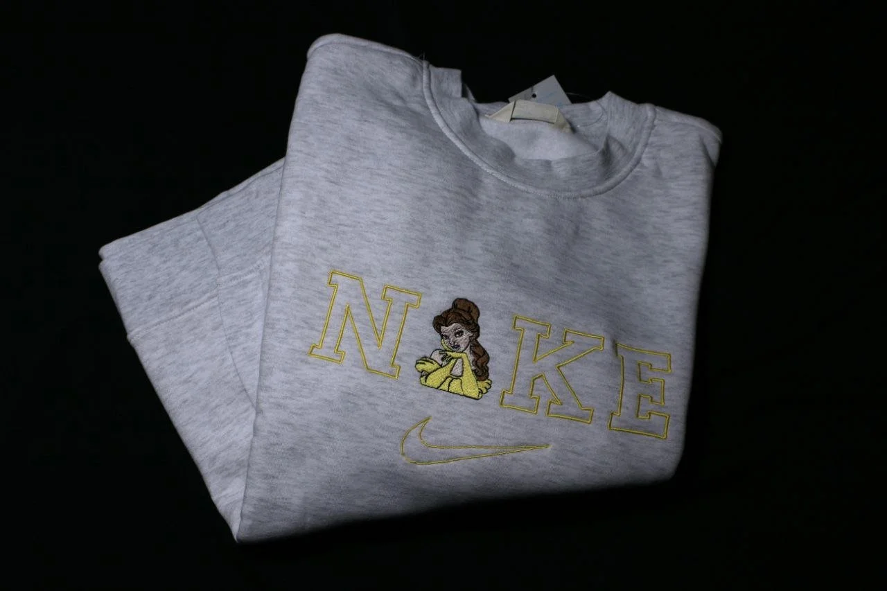 Una felpa grigia con un disegno di Belle, la protagonista di La Bella e La Bestia, sopra le lettere N e K, e il logo Nike sotto.