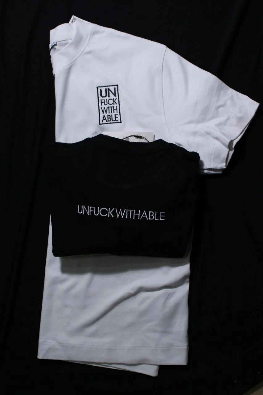 Due magliette, una bianca e una nera, con la scritta 'UNFUCKWITHABLE' sugli appartenenti, adagiate su un tessuto nero.