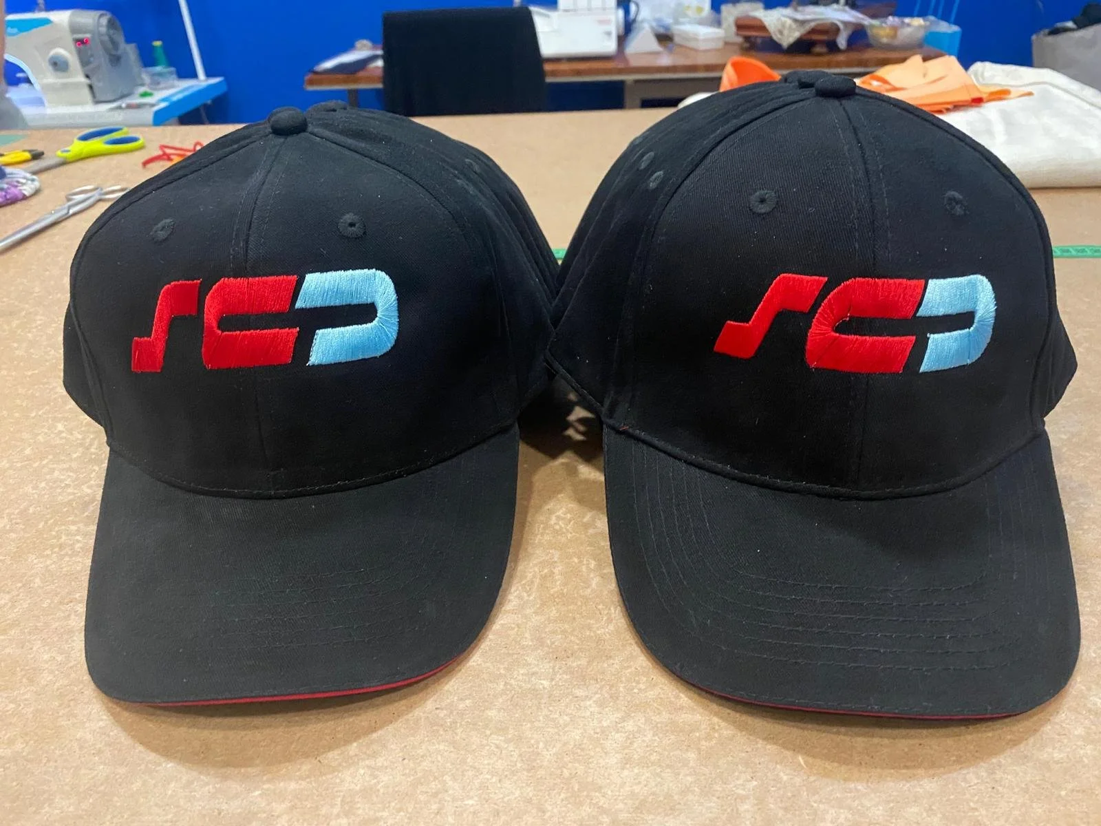 Due cappellini neri con ricamo rosso, nero e azzurro sulla fronte, posti su un tavolo di lavoro.