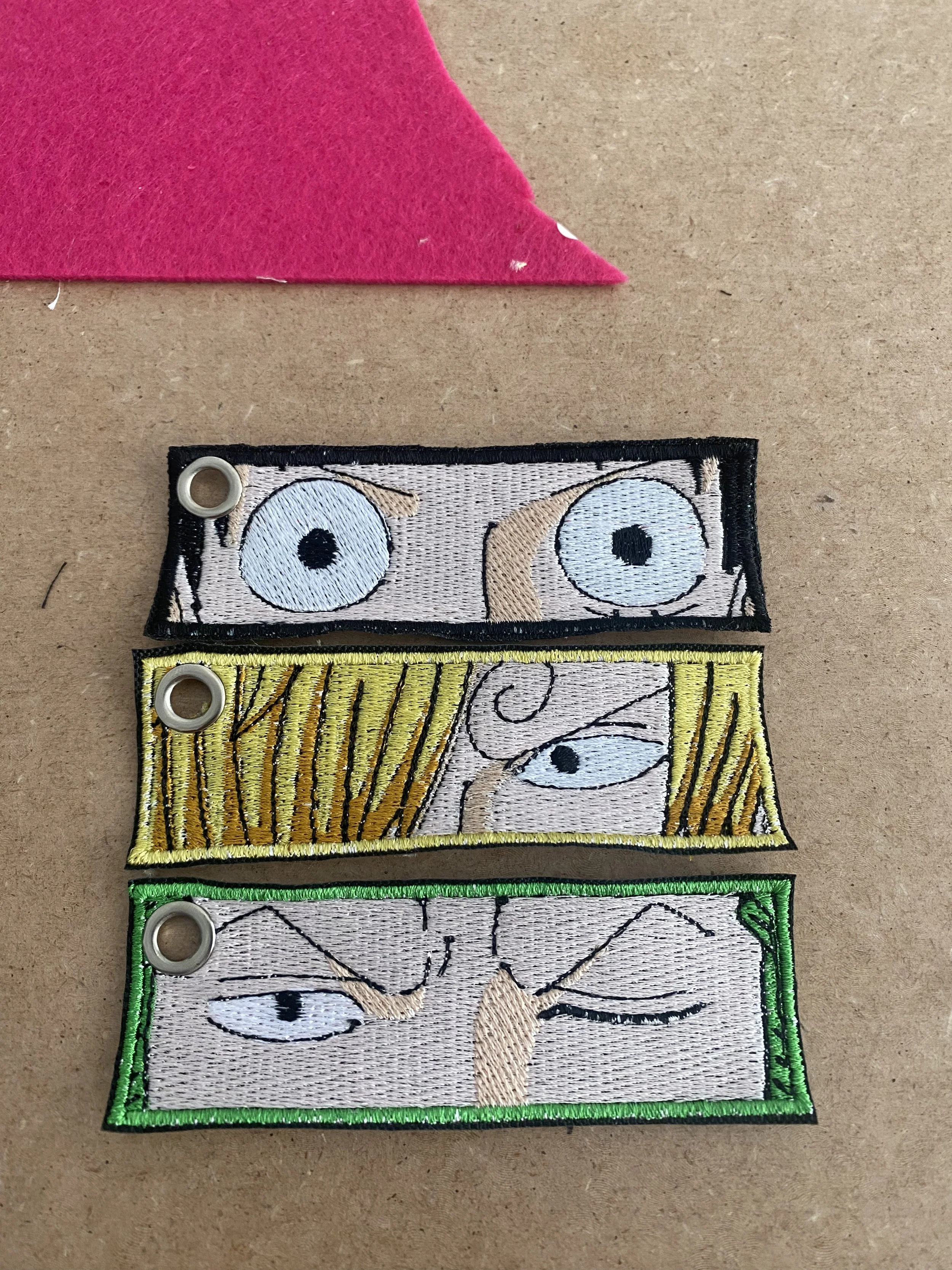 Tre toppe ricamate con dettagli di occhi e capelli di personaggi, ciascuna con occhi diversi e colori di bordatura differenti: nero, giallo, e verde.