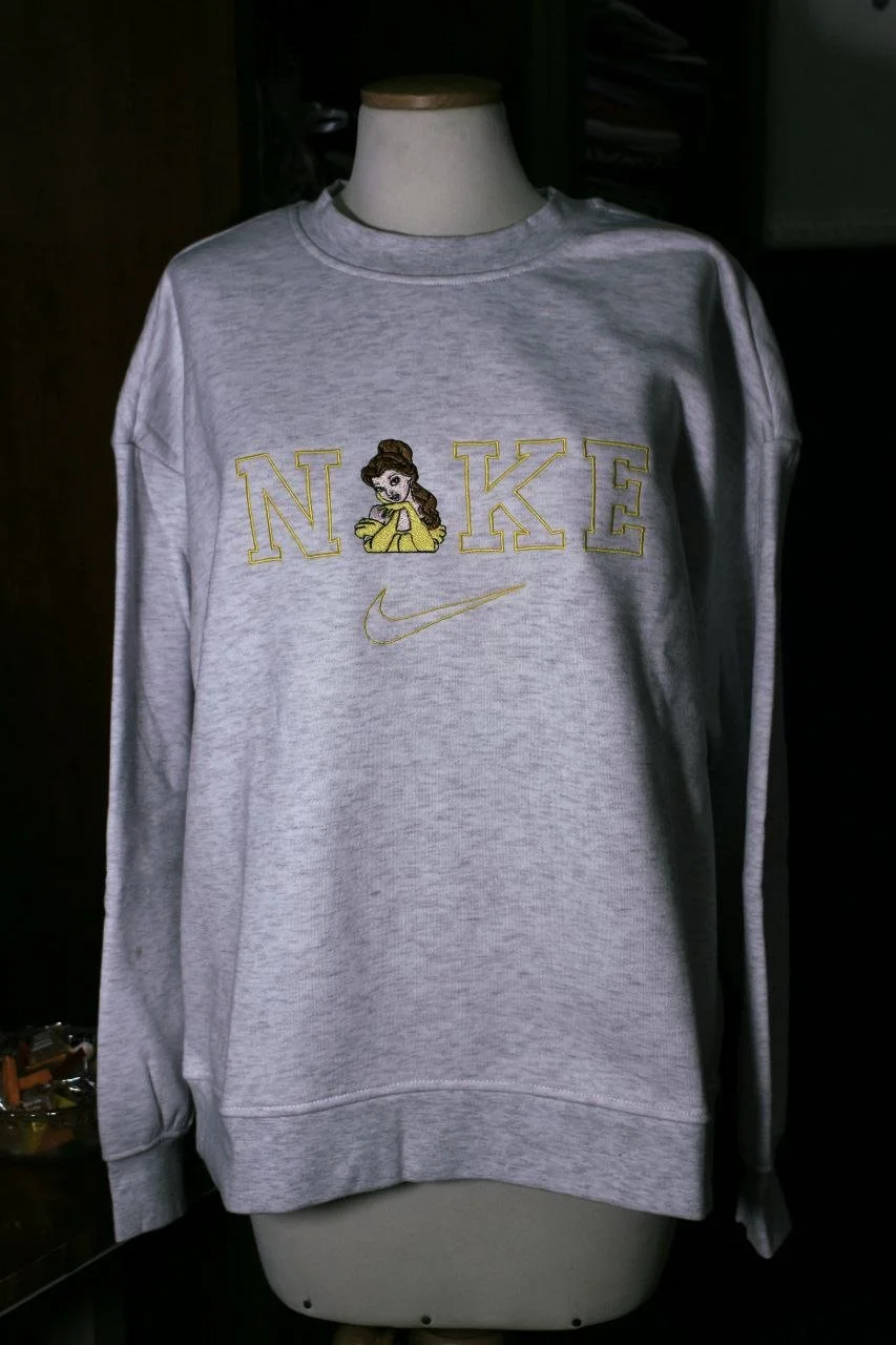 Felpa grigia con ricamo di una ragazza con cappello e l'acronimo 'NKE' in lettere gialle e il logo Nike in giallo.