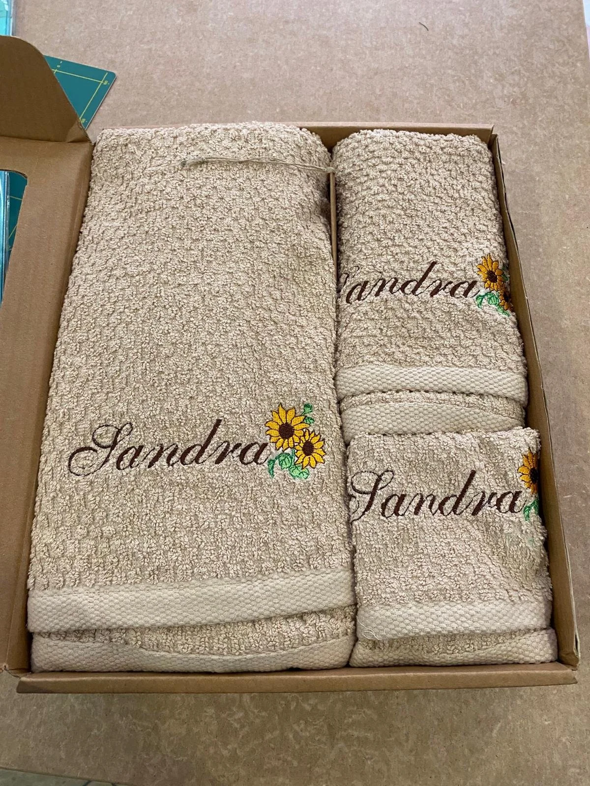 Set di asciugamani beige con ricamo della parola 'Sandra' e un disegno di girasole, impacchettati in una scatola di cartone.
