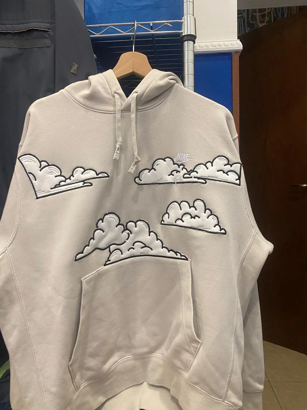 Felpa con cappuccio in tessuto chiaro, decorata con disegni di nuvole nere e bianche, firma Nike sopra le nuvole e una tasca a marsupio frontale.