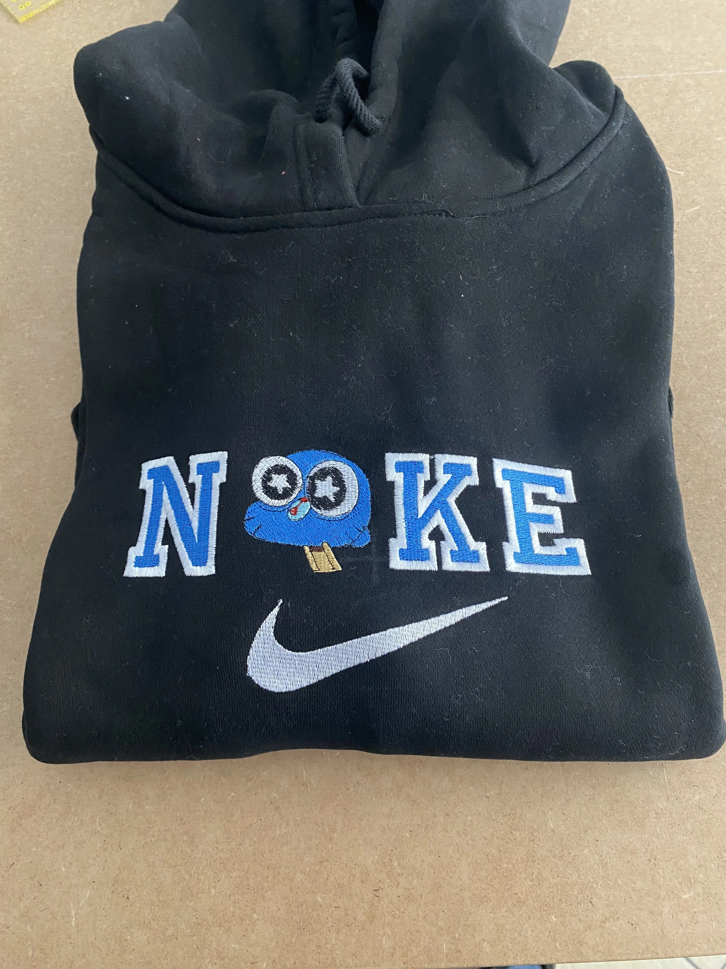 felpa nera con ricamo della parola NOKE, con una faccina stilizzata di colore blu con occhi grandi e sorriso che tiene una patente, e il logo di Nike in bianco sotto.