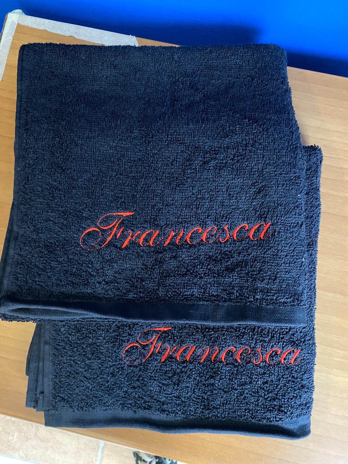 Asciugamani di colore nero con scritta rossa 'Francesca' ricamata, posizionati uno sopra l'altro su un tavolo di legno.