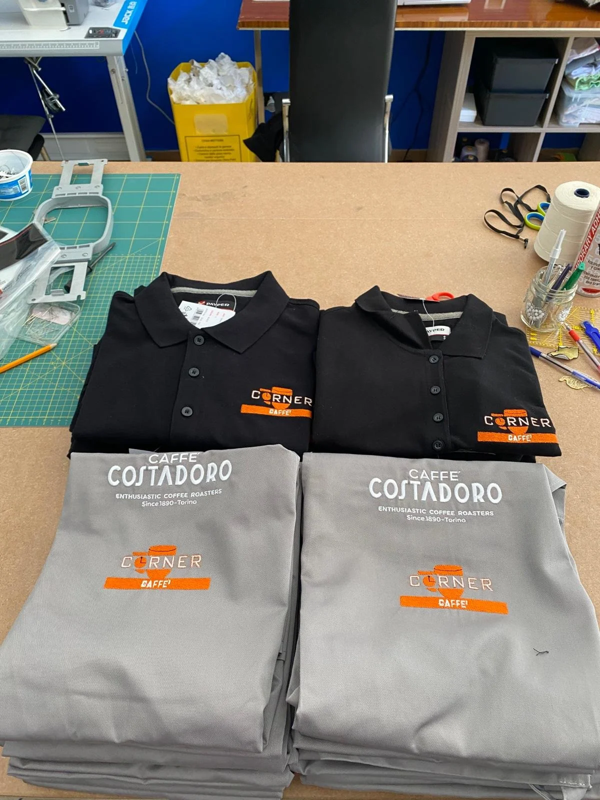 Due maniche di polo nere e quattro sacchetti grigi con logo di caffè Corner Gaffe, disposti su un banco da lavoro in un laboratorio o ufficio.