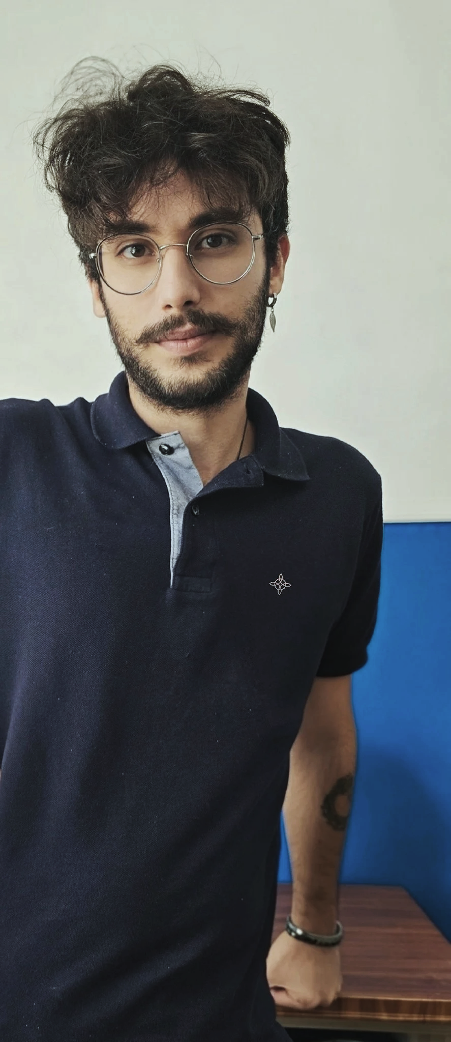 Un giovane uomo con barba, occhiali rotondi e orecchino indossa una polo nera con colletto grigio e si trova in una stanza con pareti bianche e blu, seduto davanti a un tavolo di legno.