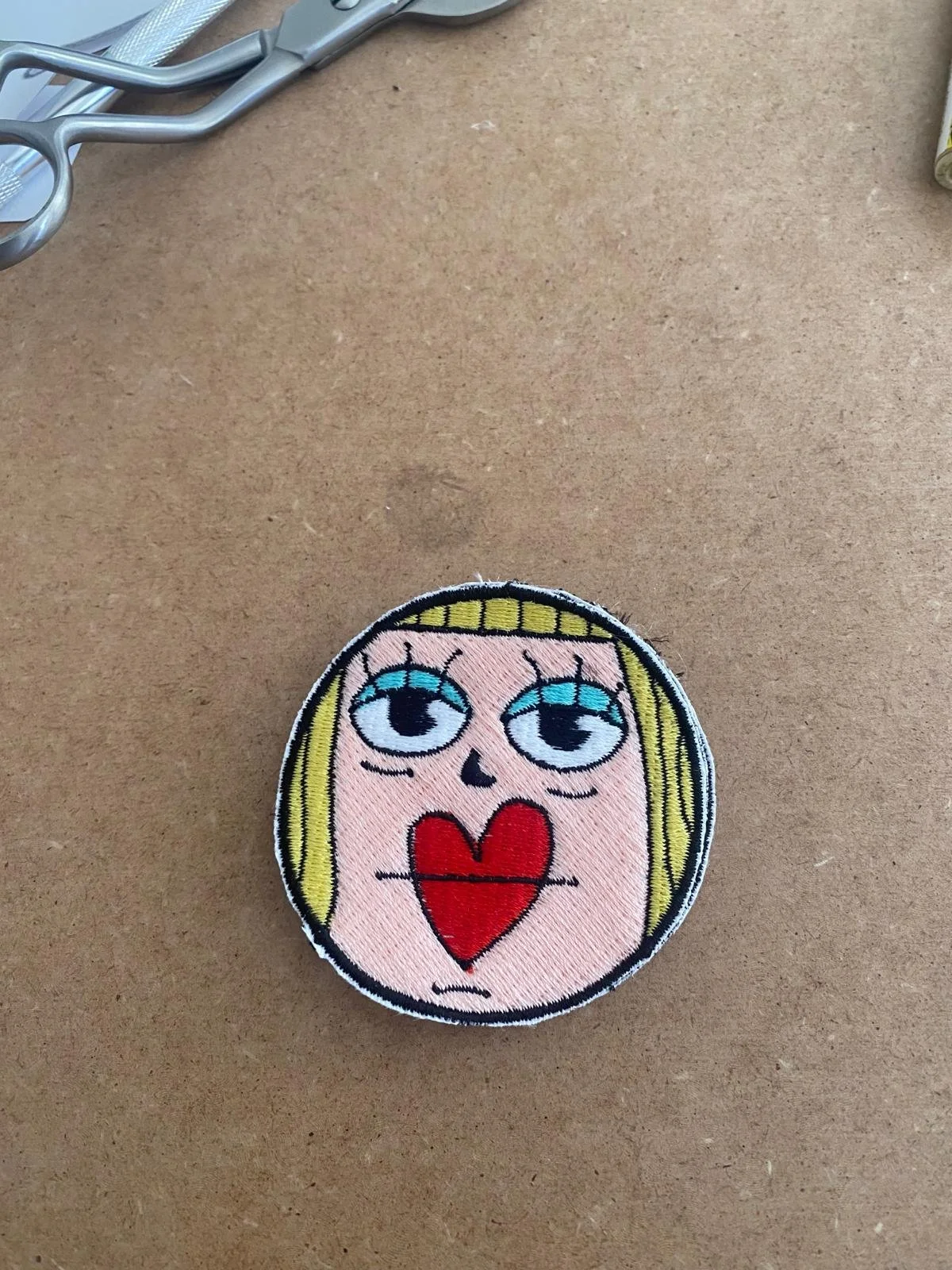 Patch ricamato con il volto di una ragazza con capelli biondi, occhi grandi, bocca con cuore rosso e espressione malinconica, su uno sfondo di colore rosa pallido.