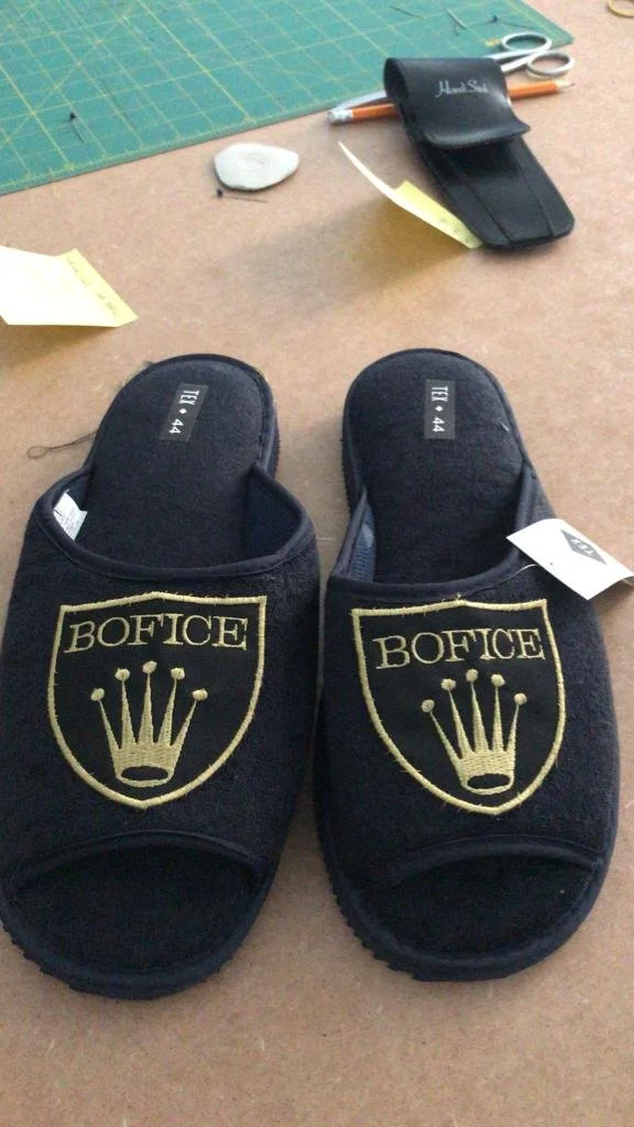 Un paio di pantofole nere con il logo 'BOFFICE' e una corona stilizzata, posizionate su una superficie di lavoro. Sullo sfondo si vedono forbici, penne, un rotolo di carta adesiva e altri oggetti da ufficio.