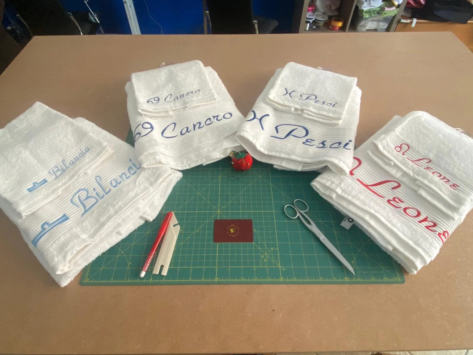 Lenzuola bianche con scritte colorate, disposte su un tavolo, con strumenti di cucito e un tappetino da taglio al centro.