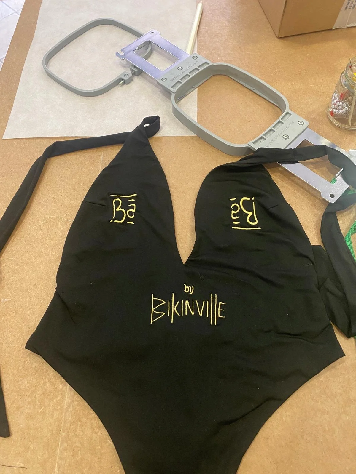 Un cappellino da cosmonautica nero con scritte gialle che recitono "Bébé by BikiniVJE"; sopra un tavolo si trovano una struttura metallica e un telaio da cucire.