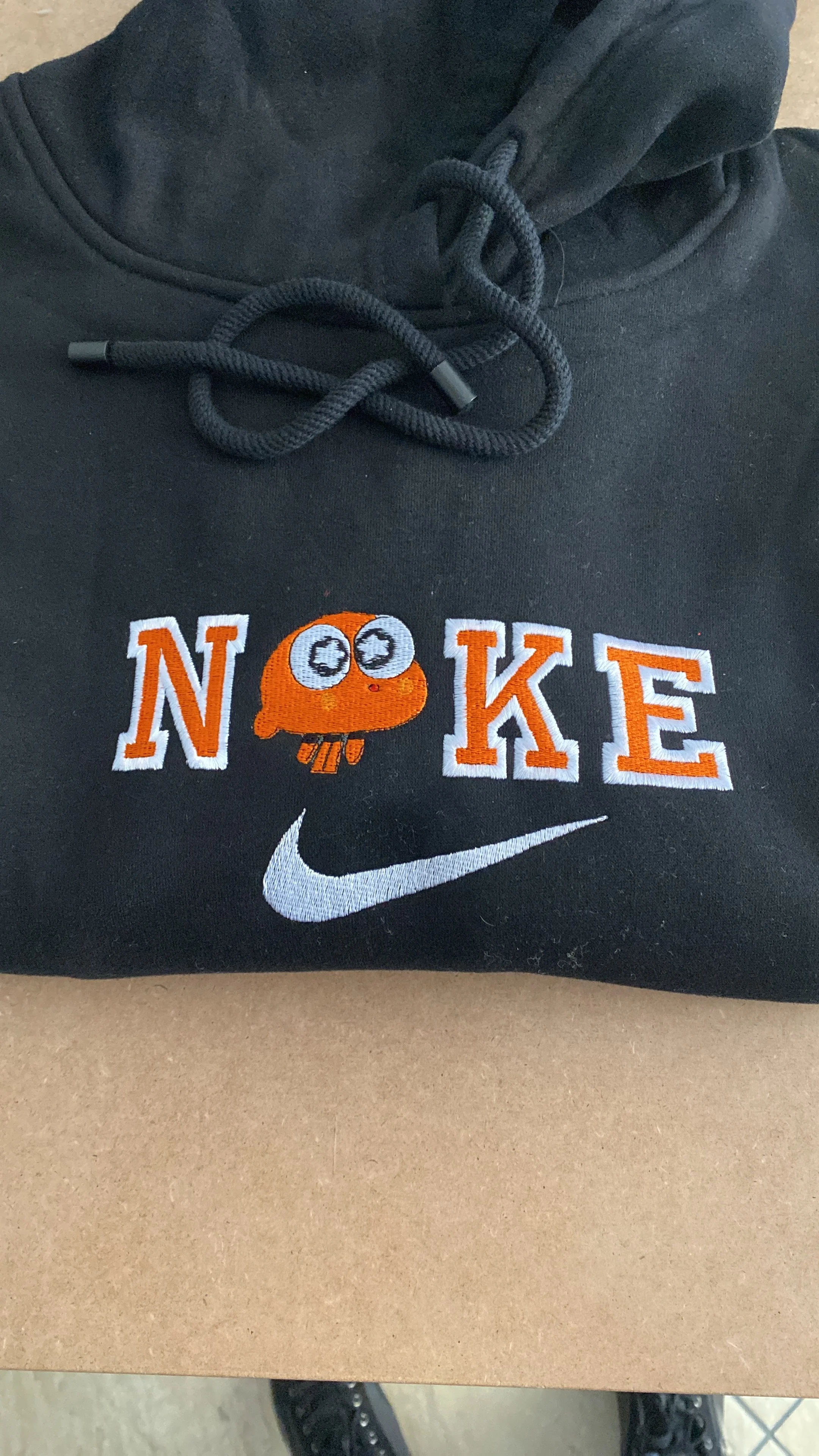 Felpa nera con cappuccio con ricamo di una mascotte di un calamaro arancione con occhi grandi e il logo Nike sotto.