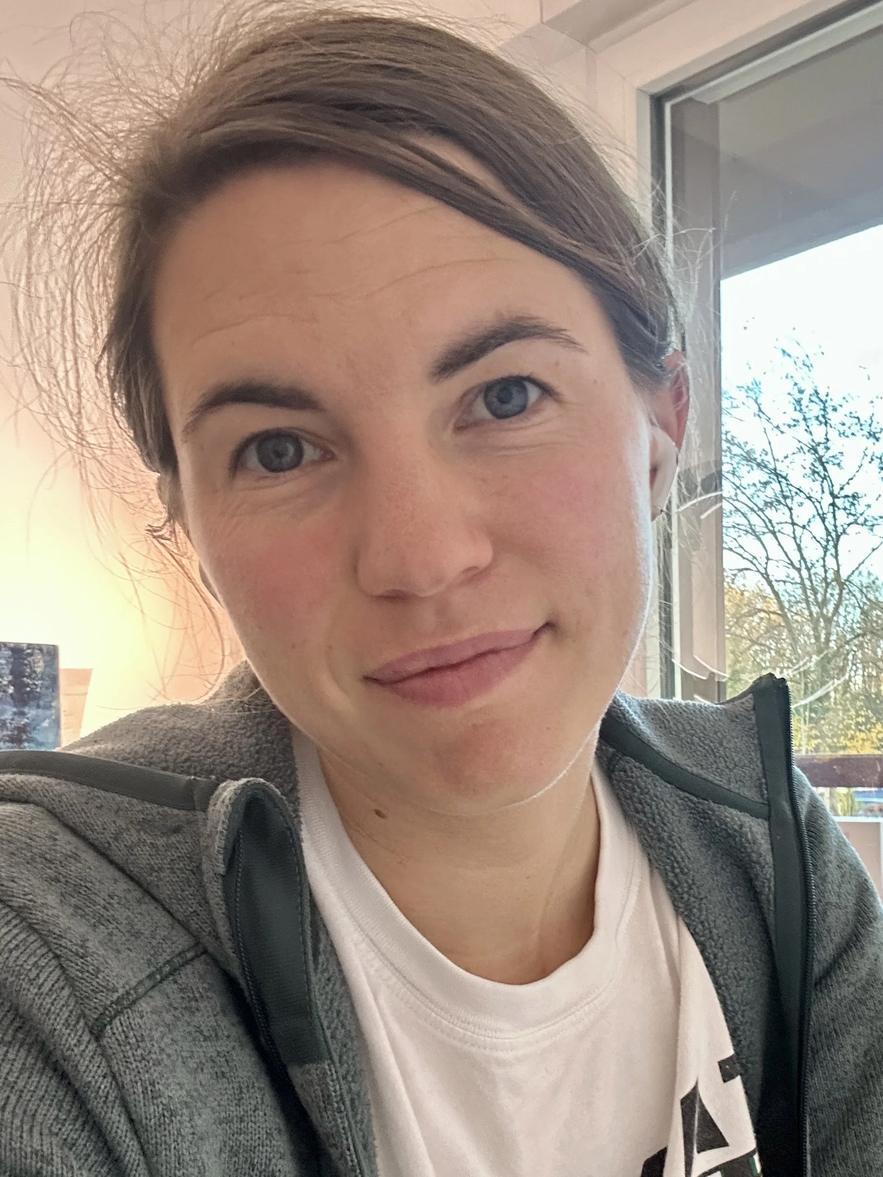 Nahaufnahme einer jungen Frau mit braunen Haaren als Zopf zusammengefasst, blauen Augen, in einem hell erleuchteten Raum, mit Fenster im Hintergrund und einem Baum draußen.