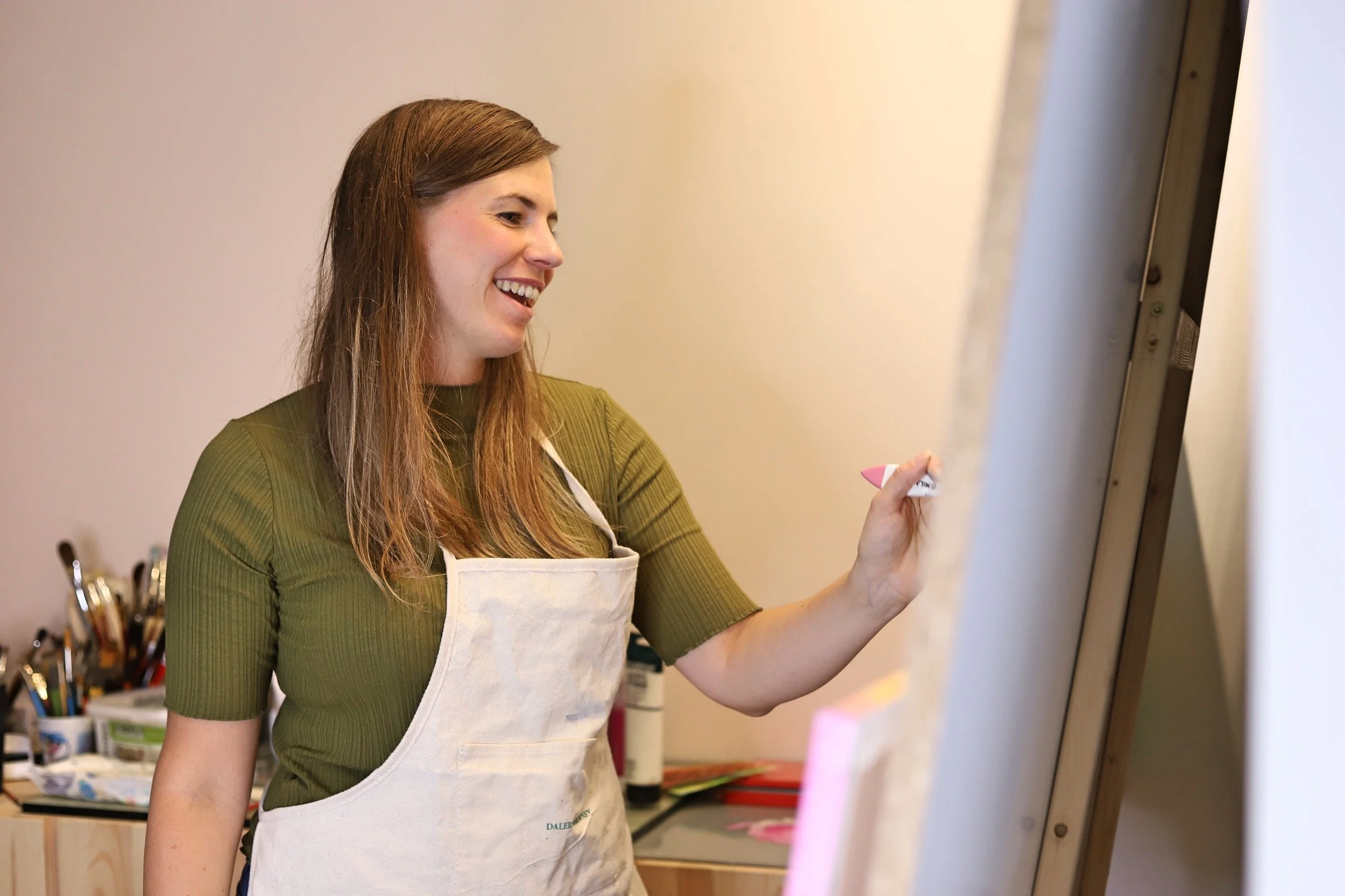 Laura Franz malt auf einer Staffelei in einem Kunststudio, trägt einen grünes T-Shirt und eine weiße Schürze und lächelt während sie malt.