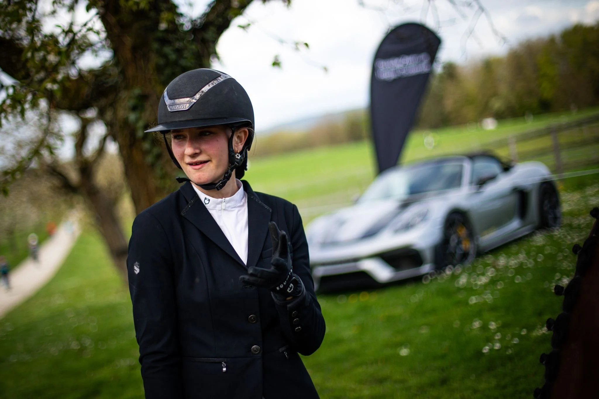 Une femme portant un casque de cheval et des vêtements d'équitation venant du site internet EponaStore et de la marque Equestro, en plein air, avec une voiture de sport Porsche gris foncé en arrière-plan. Les vêtements sont de qualités, sobres et uni
