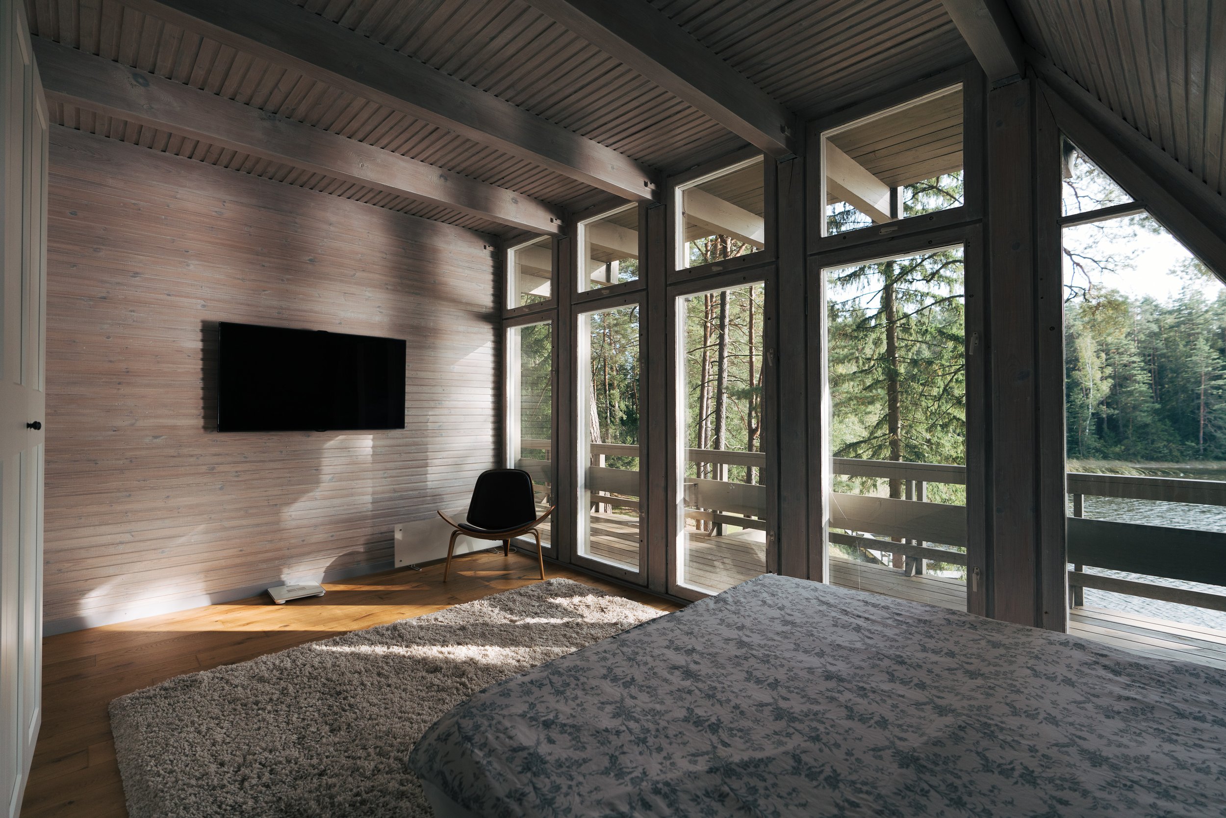 Room with large glass windows overlooking a forest, with a bed, a black chair, a TV mounted on a wooden wall, and a beige rug.