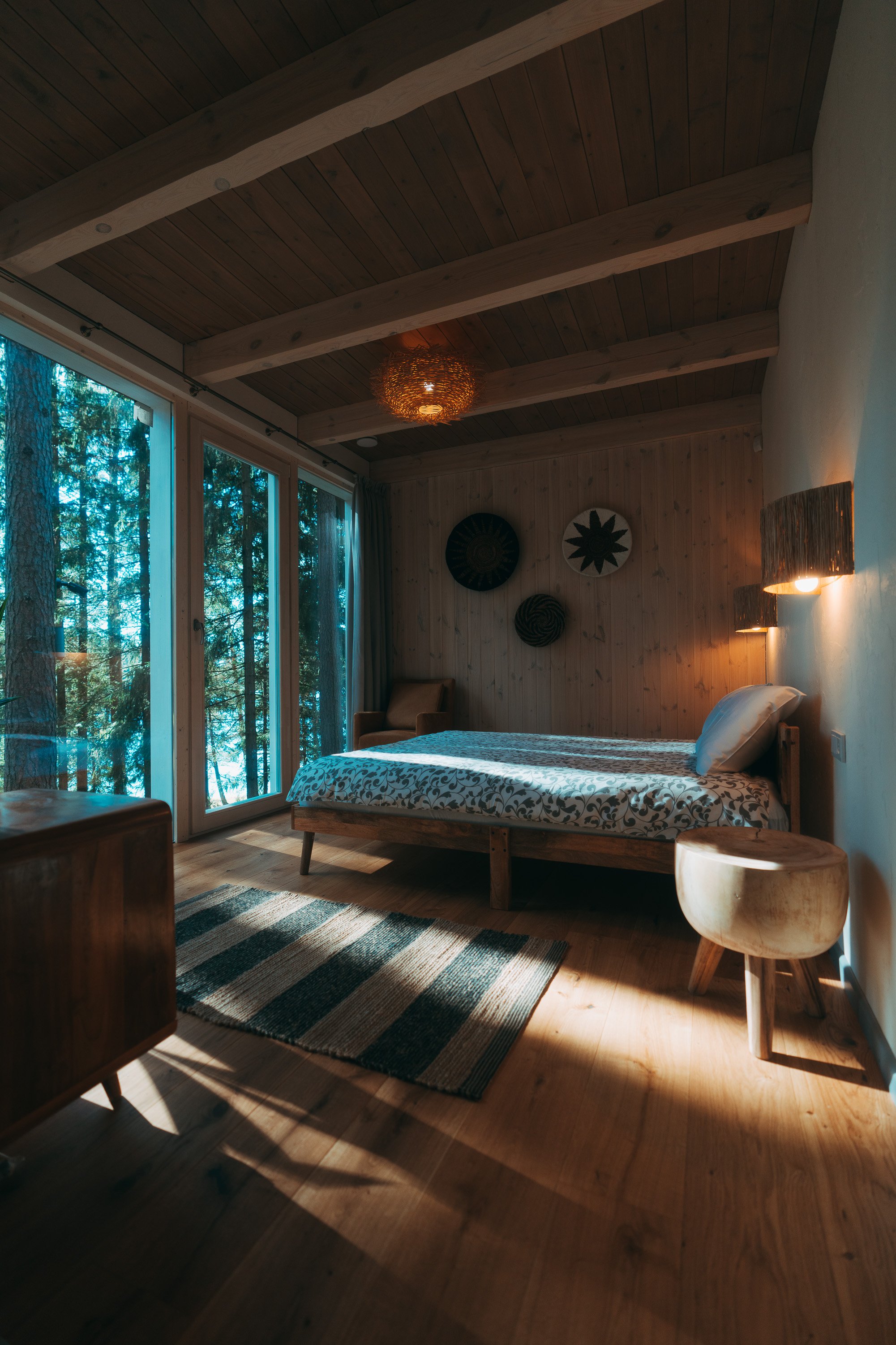 A cozy bedroom with wooden walls and ceiling, a bed with patterned bedding, a small round wooden stool, an armchair by large windows showing a forest outside, and wall-mounted lamps.