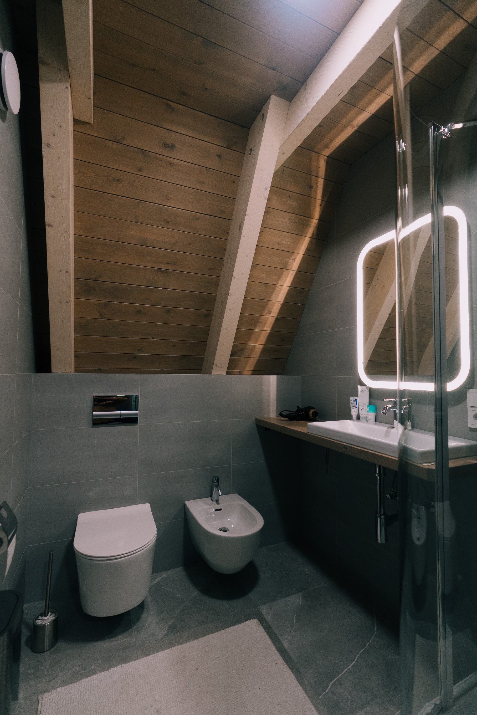 Modern bathroom with sloped wooden ceiling, green tiled walls, white toilet and bidet, wall-mounted sink with illuminated mirror, gray floor tiles, and a white rug.