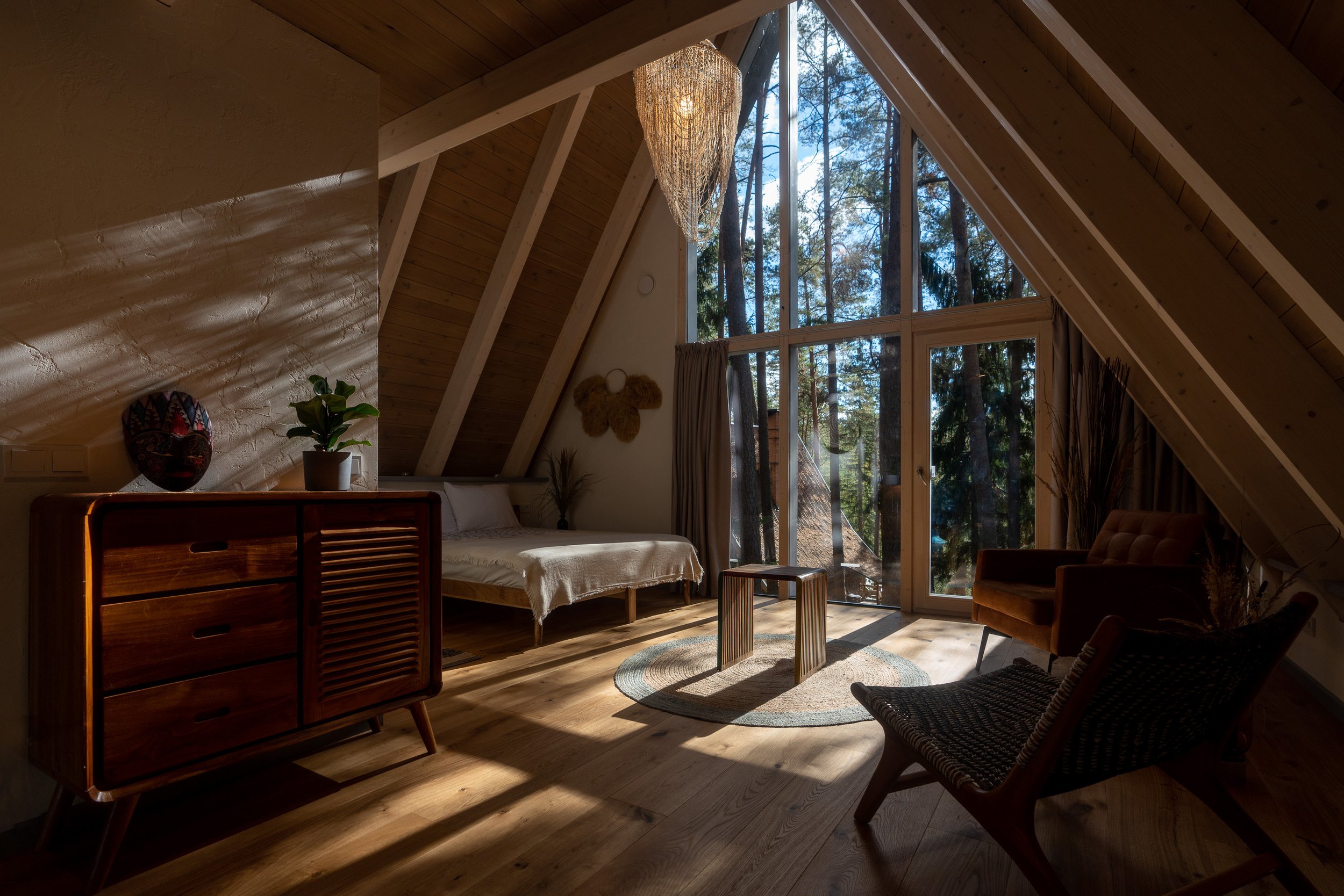Interior of a cozy attic bedroom with wooden walls, large windows showing tall trees, a bed with white bedding, a small wooden table, two chairs, a dresser, and decorative plants and artwork.