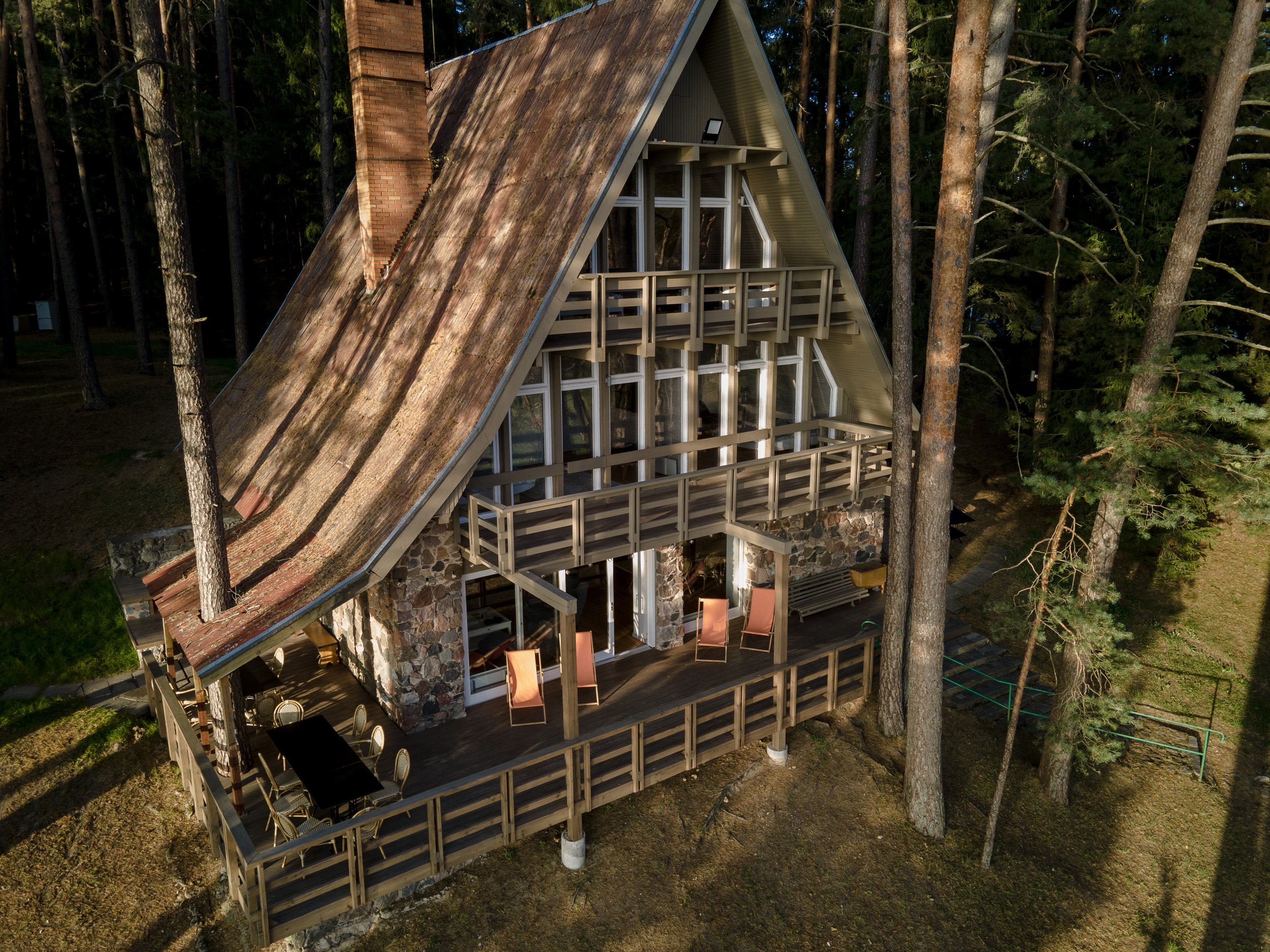 A tall A-frame house with a large deck surrounded by trees, featuring a stone base, glass windows, and outdoor furniture including chairs and a dining table.