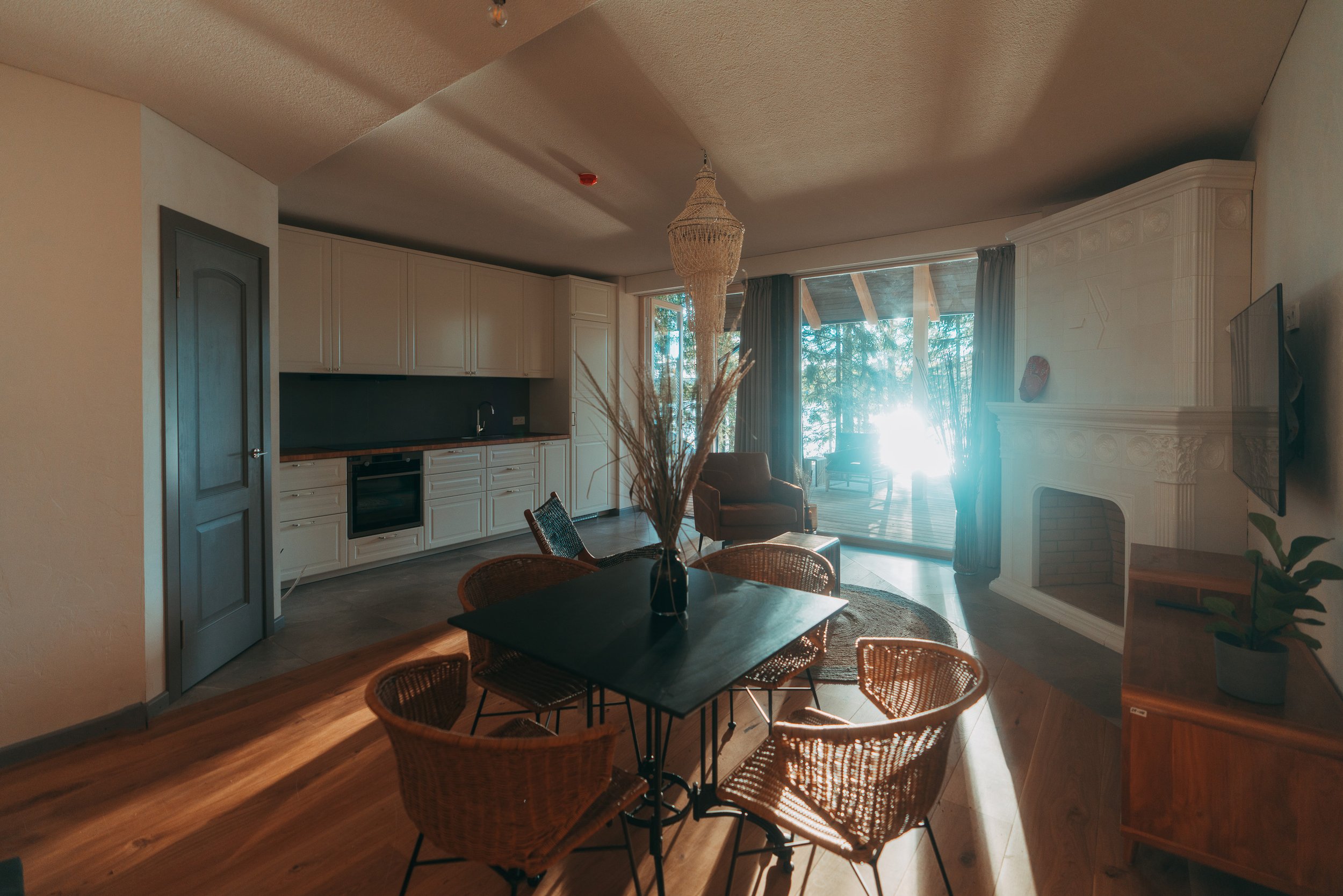 Living room with a dining table and chairs, a white fireplace, large sliding glass door, and kitchen area in the background. Bright sunlight streaming through the windows.