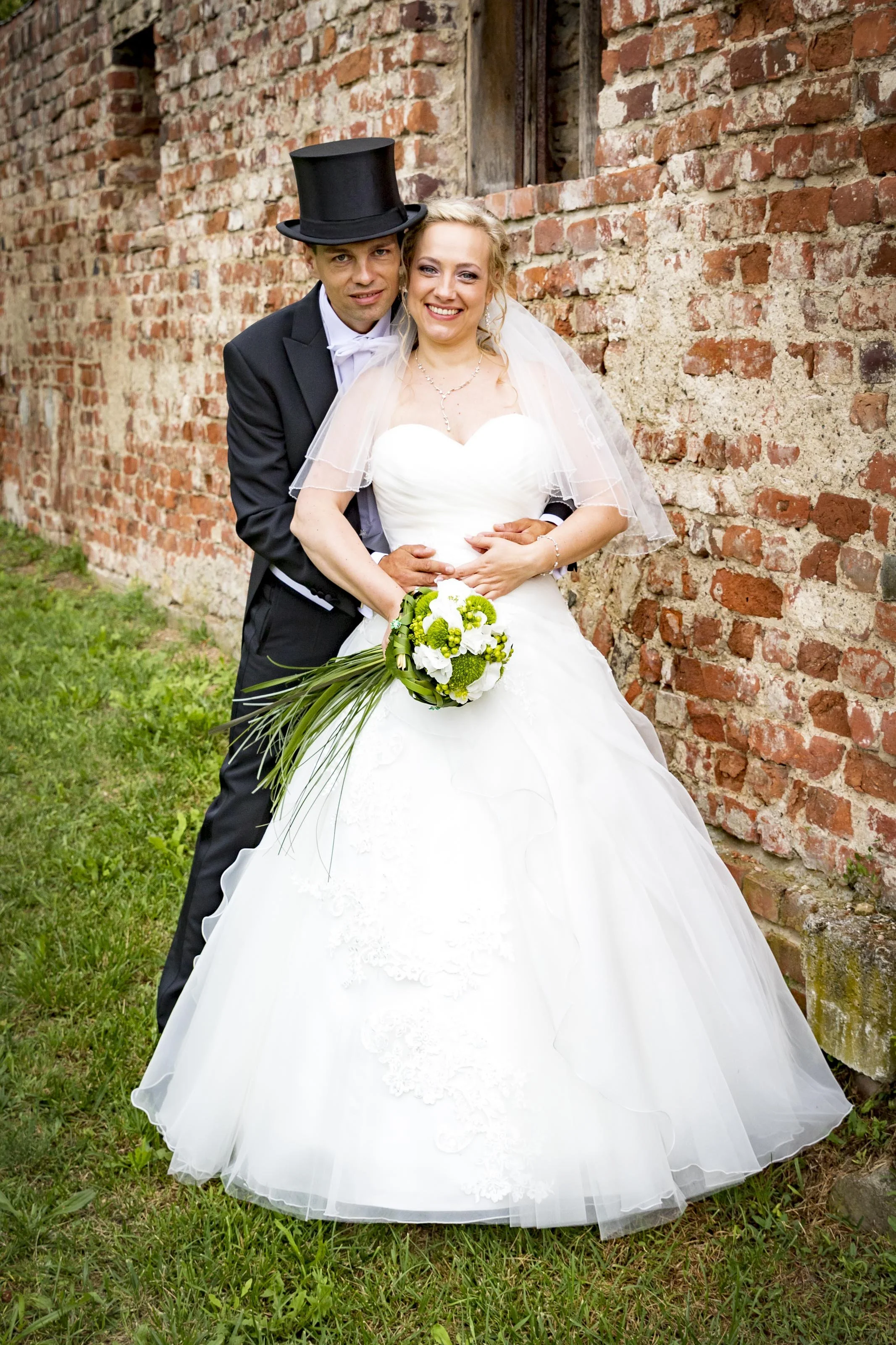 Verheiratetes Paar in Hochzeitskleidung vor einer alten Backsteinmauer, lachend und umarmend.