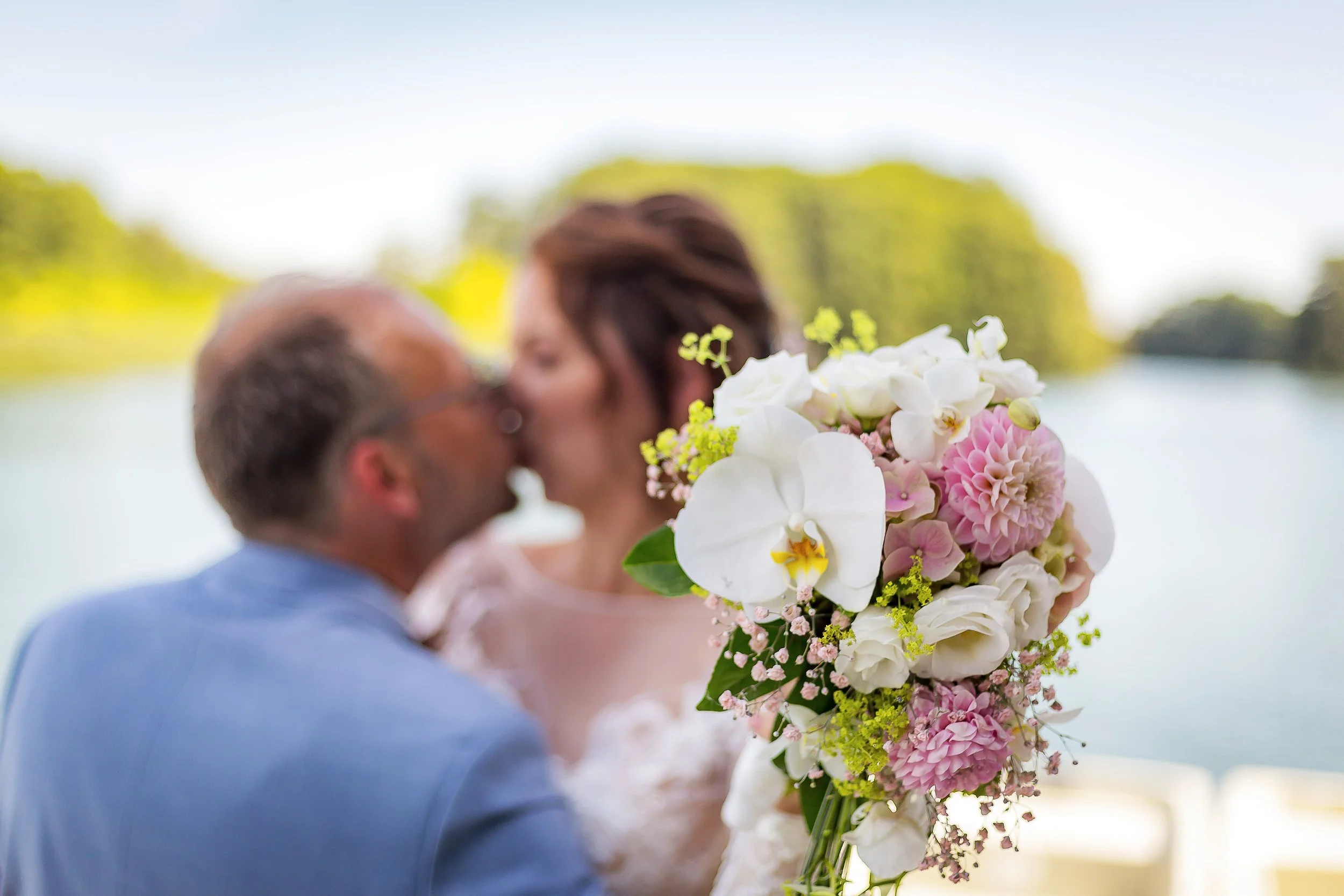 Ein Brautpaar küsst sich mit Blick auf einen Fluss im Hintergrund, während die Braut ein großes, farbenfrohes Blumenbouquet hält.