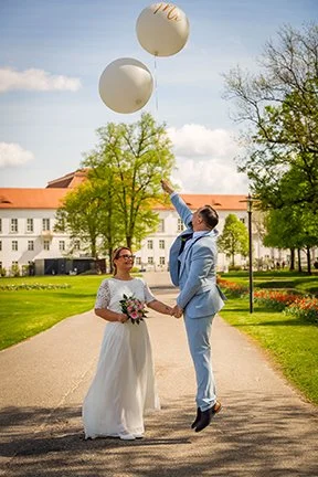 Ein Brautpaar hält die Hände und lässt drei weiße Ballons in den Himmel steigen, im Park bei sonnigem Himmel und Bäumen.