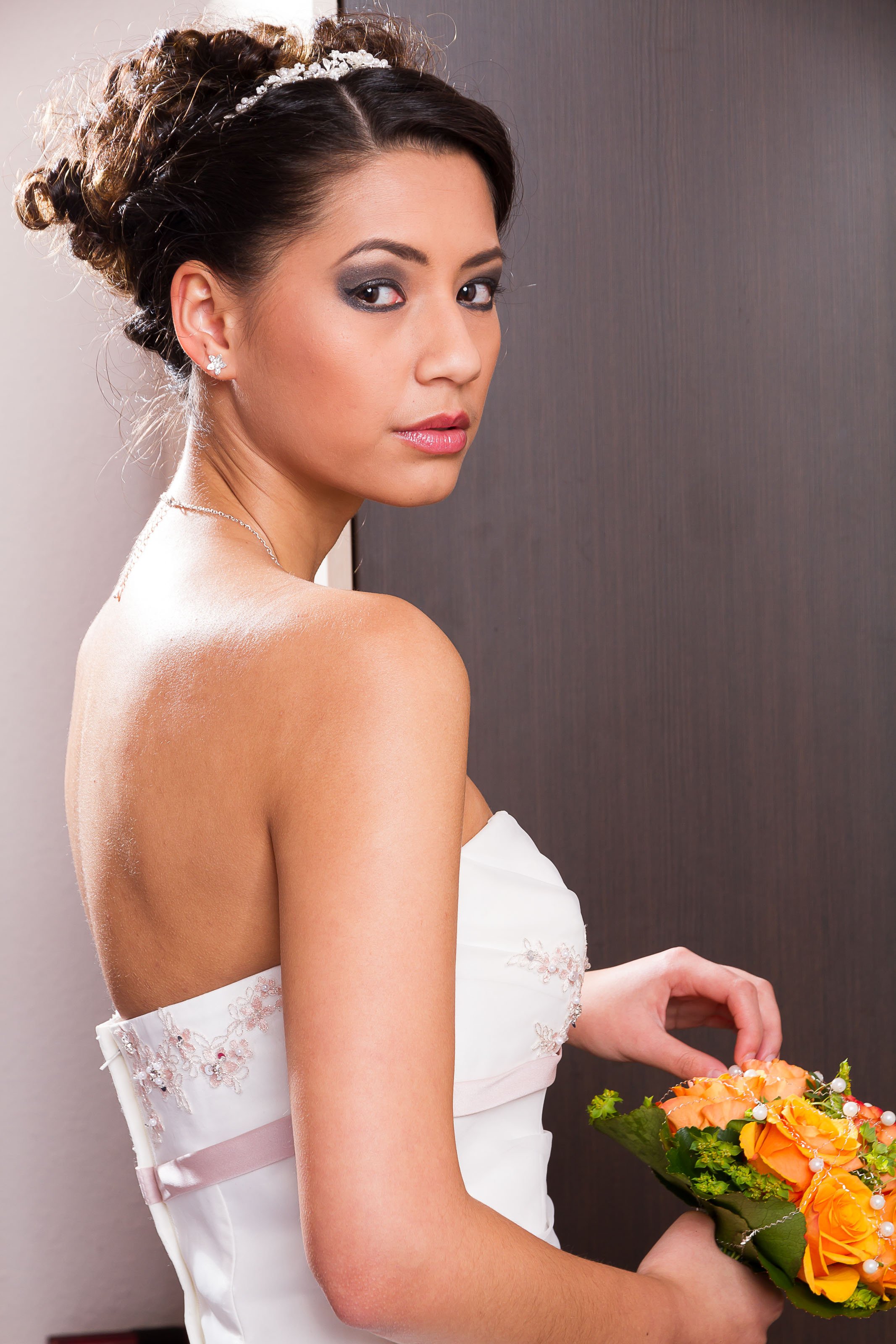 Junge Frau in Hochzeitskleid mit Brautstrauß, dunkle, lockige Haare, auffälliges Make-up, im Hintergrund eine dunkle Wand.