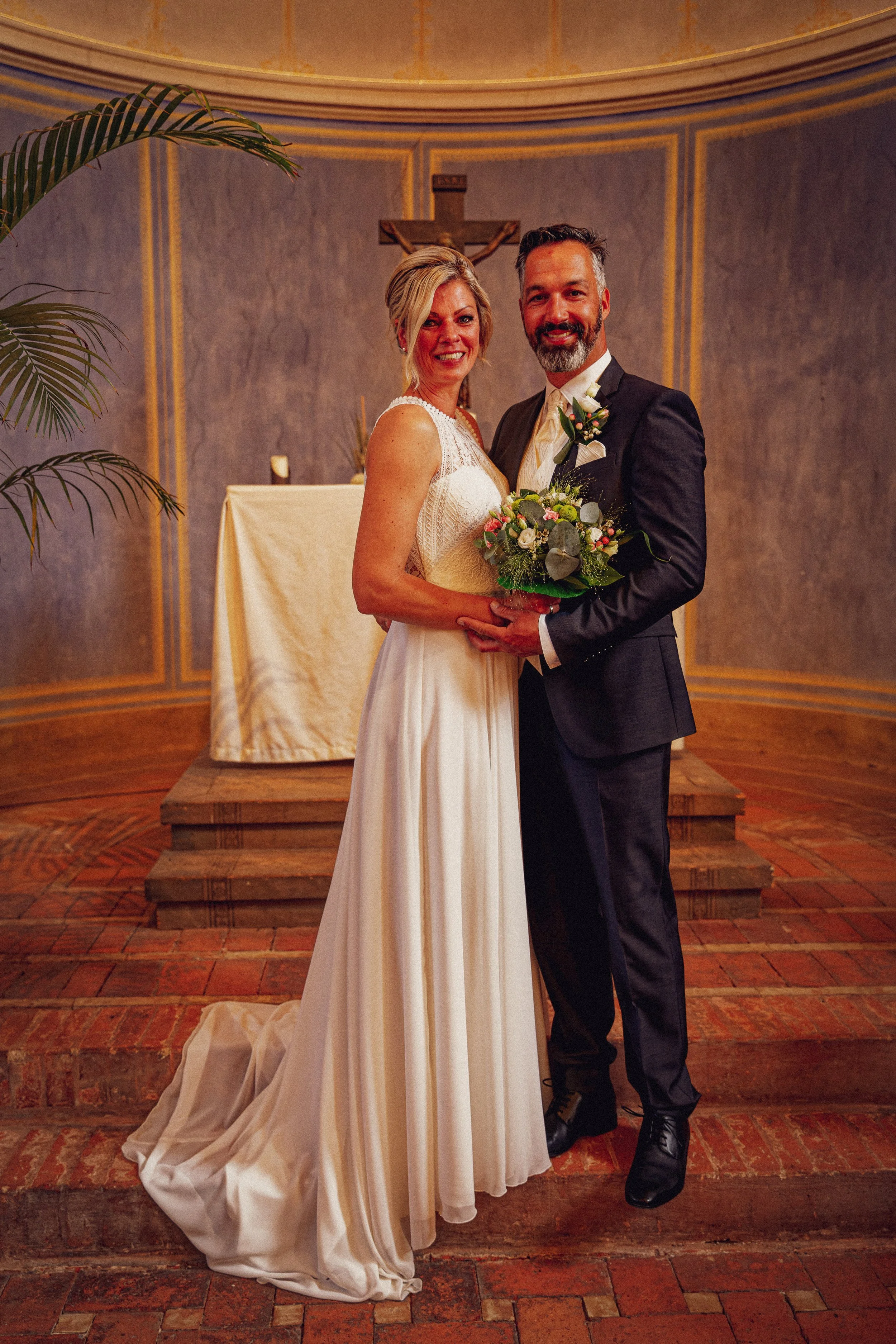 Braut und Bräutigam stehen sich in einer Kirche, die Braut trägt ein langes weißes Kleid, der Bräutigam ein schwarzes Sakko, beide halten einen Blumenstrauß, im Hintergrund ein Kreuz und eine Altar