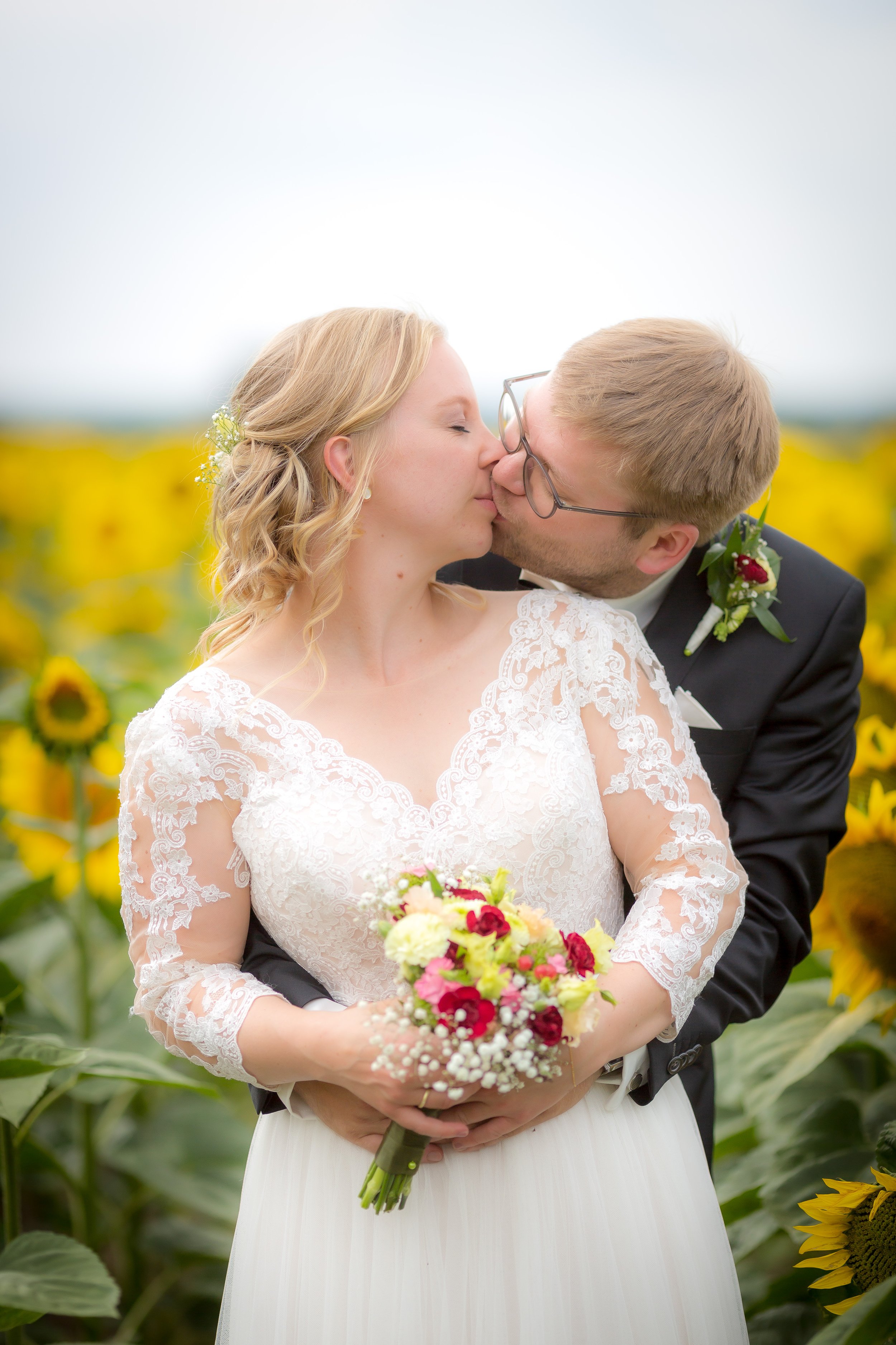 Ein Brautpaar küsst sich in einem Sonnenblumenfeld, die Braut trägt ein weißes Spitzenkleid und hält einen Blumenstrauß, der Bräutigam trägt einen schwarzen Anzug mit Krawatte.