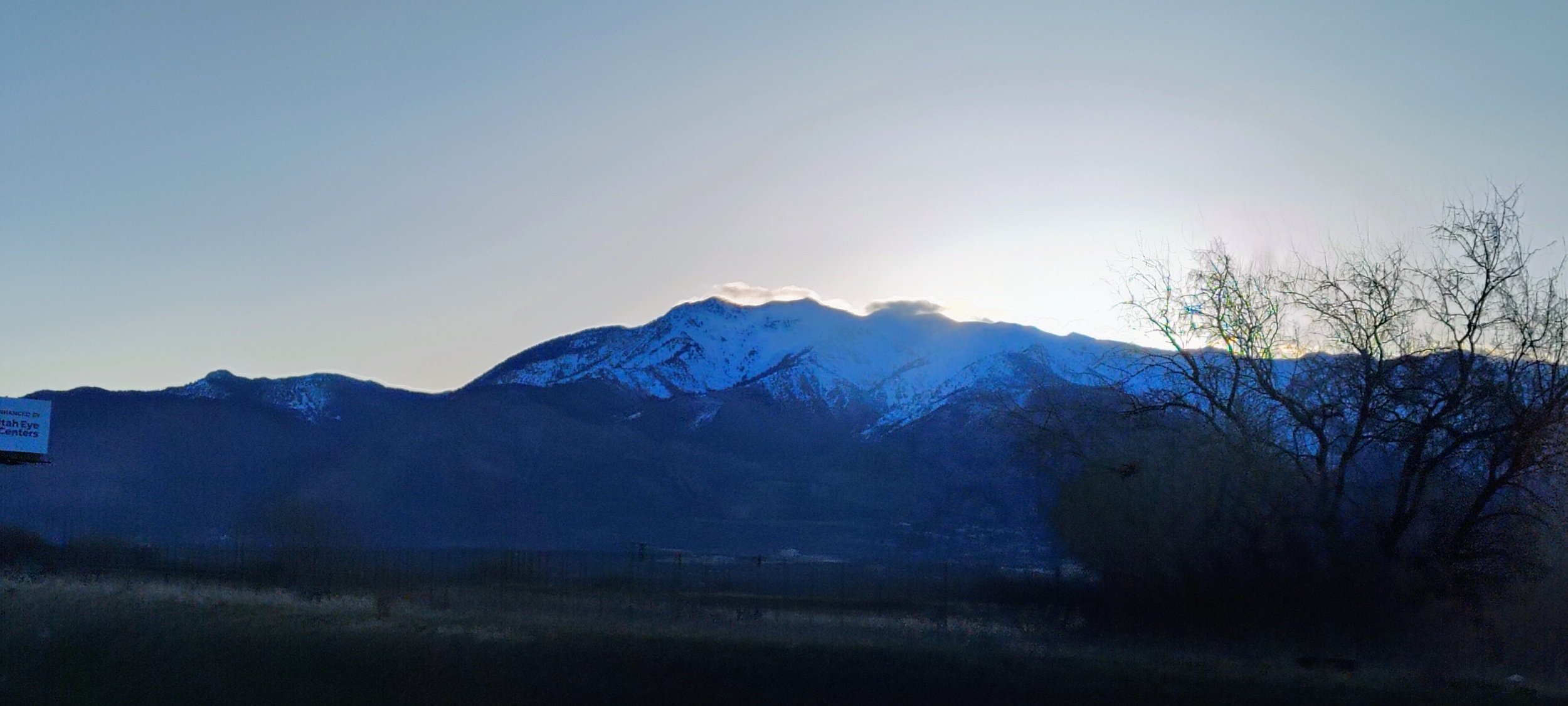 Snow-capped mountain range with a clear sky at sunset or sunrise, minimal trees in the foreground.