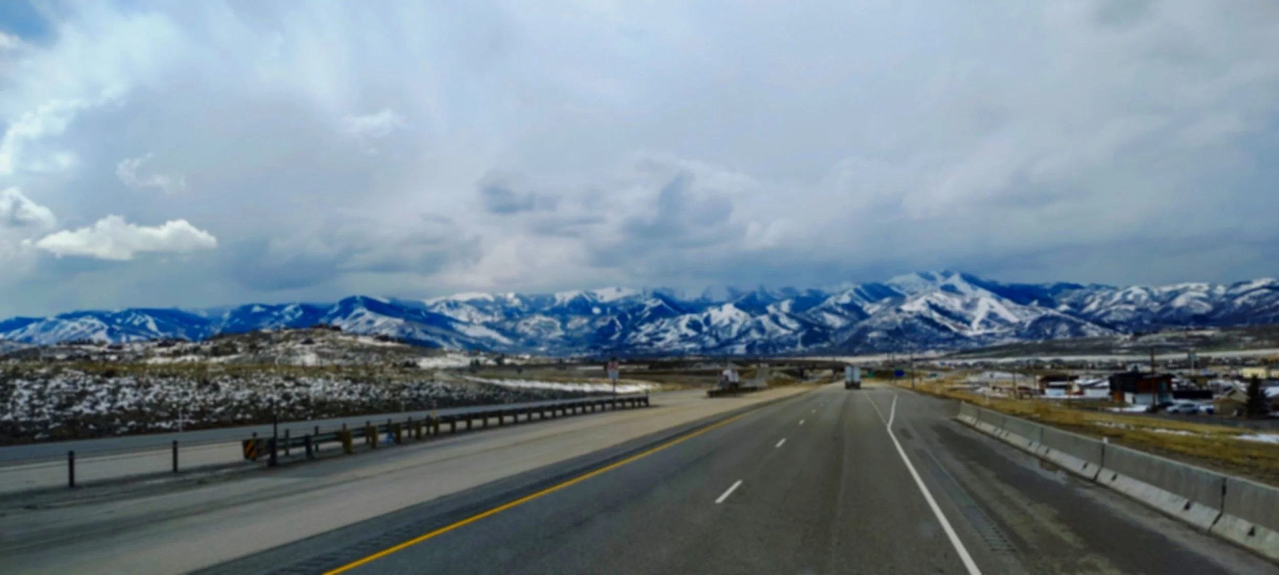 A highway with mountains covered in snow in the background and a cloudy sky overhead.