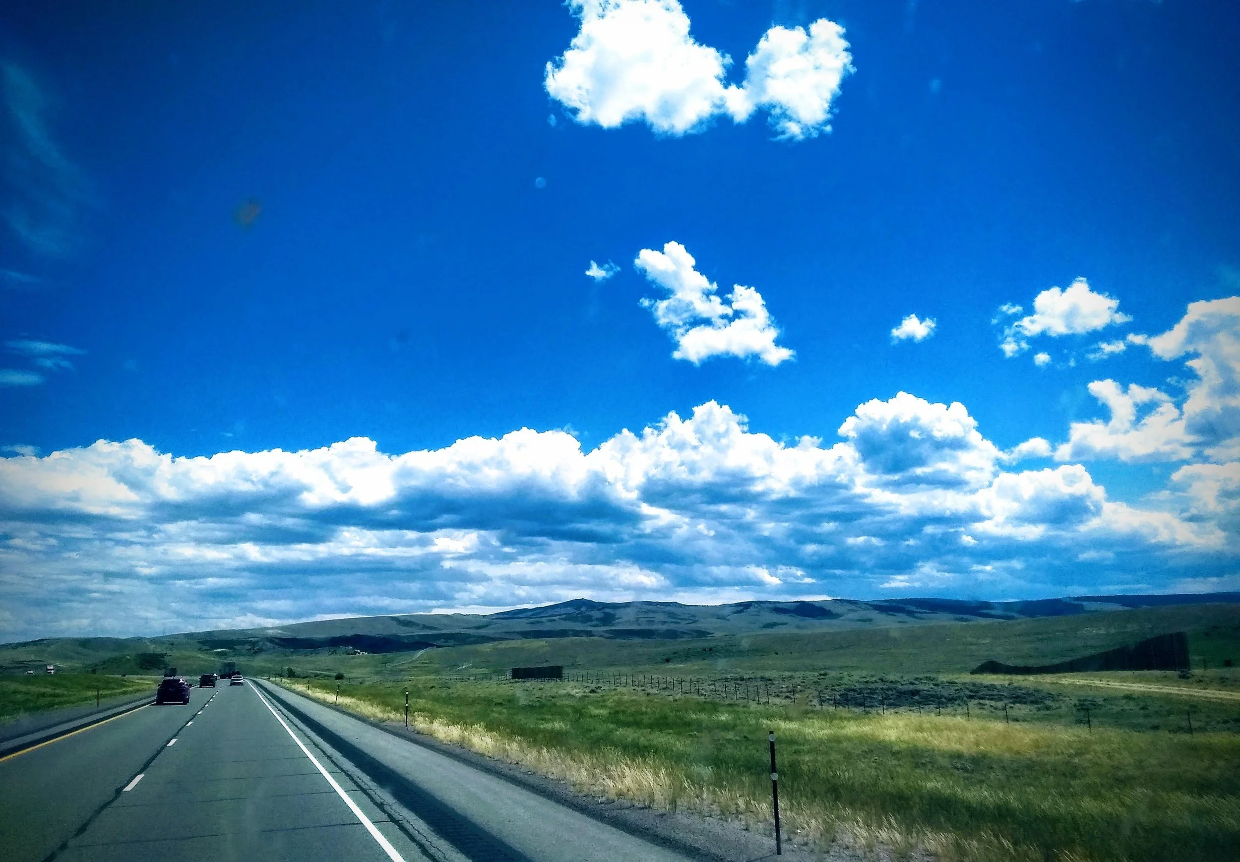 A highway running through green rolling hills under a bright blue sky with scattered fluffy white clouds.