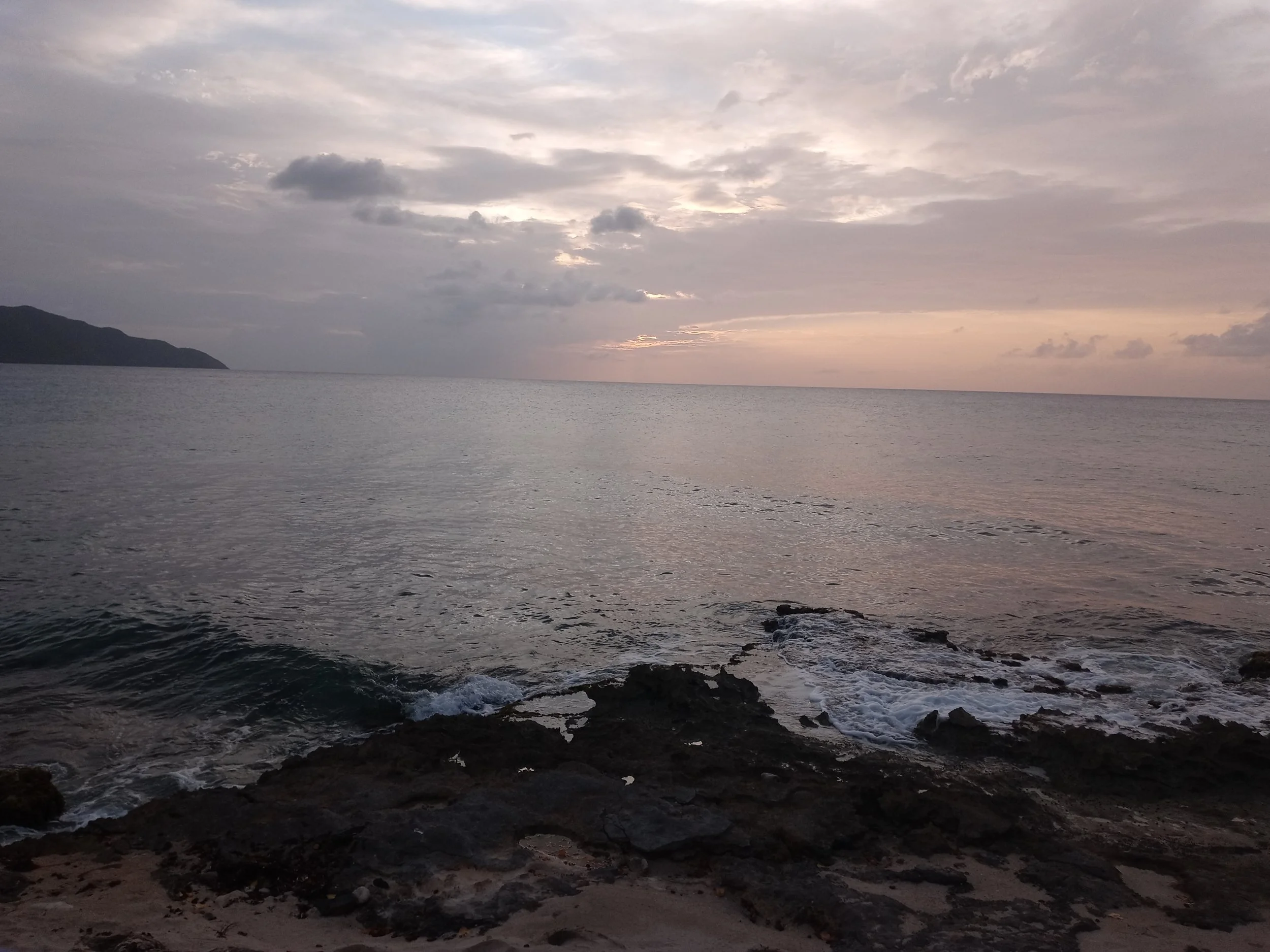 A sunset over the ocean with cloudy sky, distant land on the left, rocky shore in the foreground, gentle waves