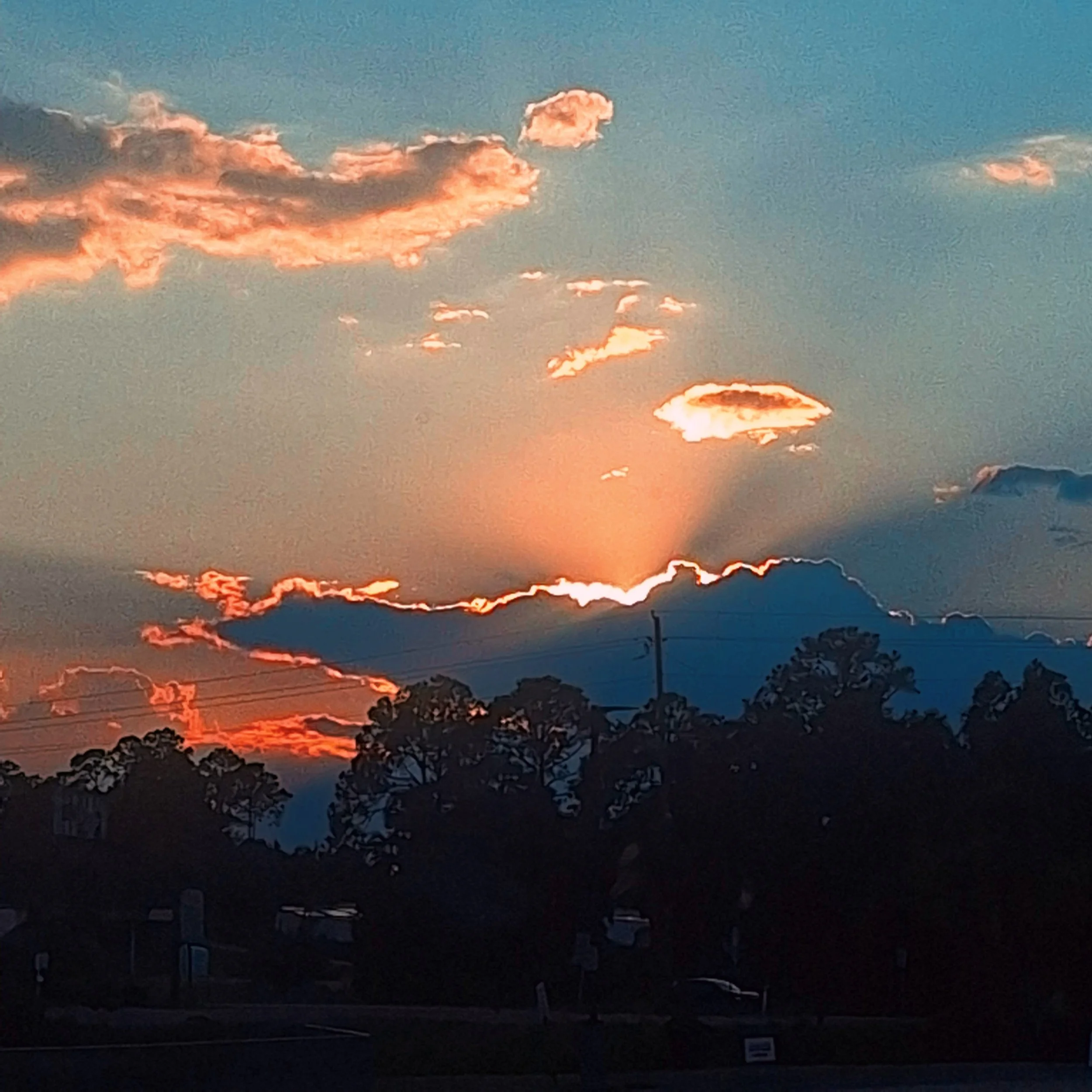 Sunset with vibrant orange and pink hues behind dark clouds over a silhouette of trees and power lines in the foreground.
