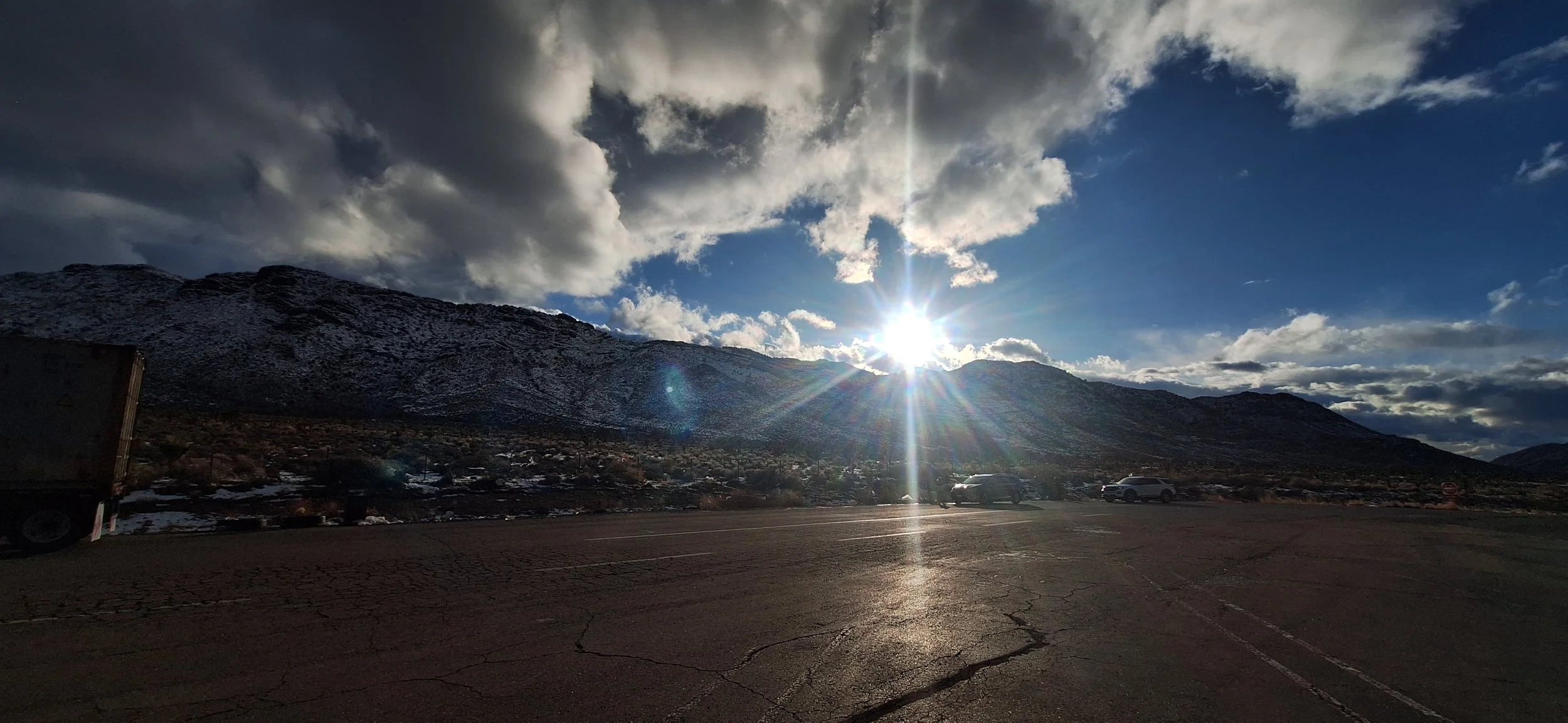 A parking lot with a mountain range in the background, snow-covered peaks, and a bright sun shining through scattered clouds in the sky.