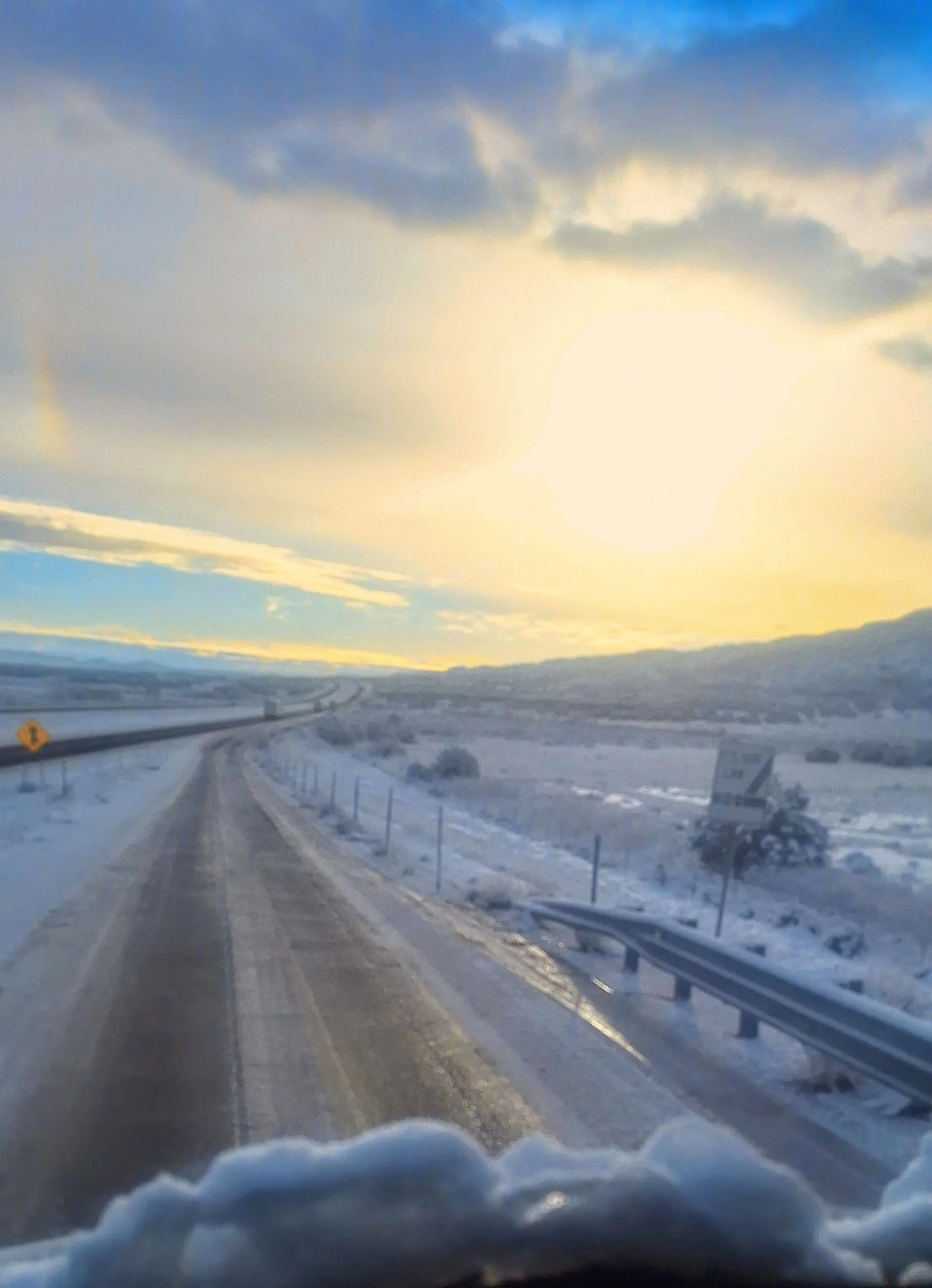 A snow-covered highway with guardrails on the right side, leading into a landscape of snow and hills under a bright sky with the sun partially obscured by clouds.