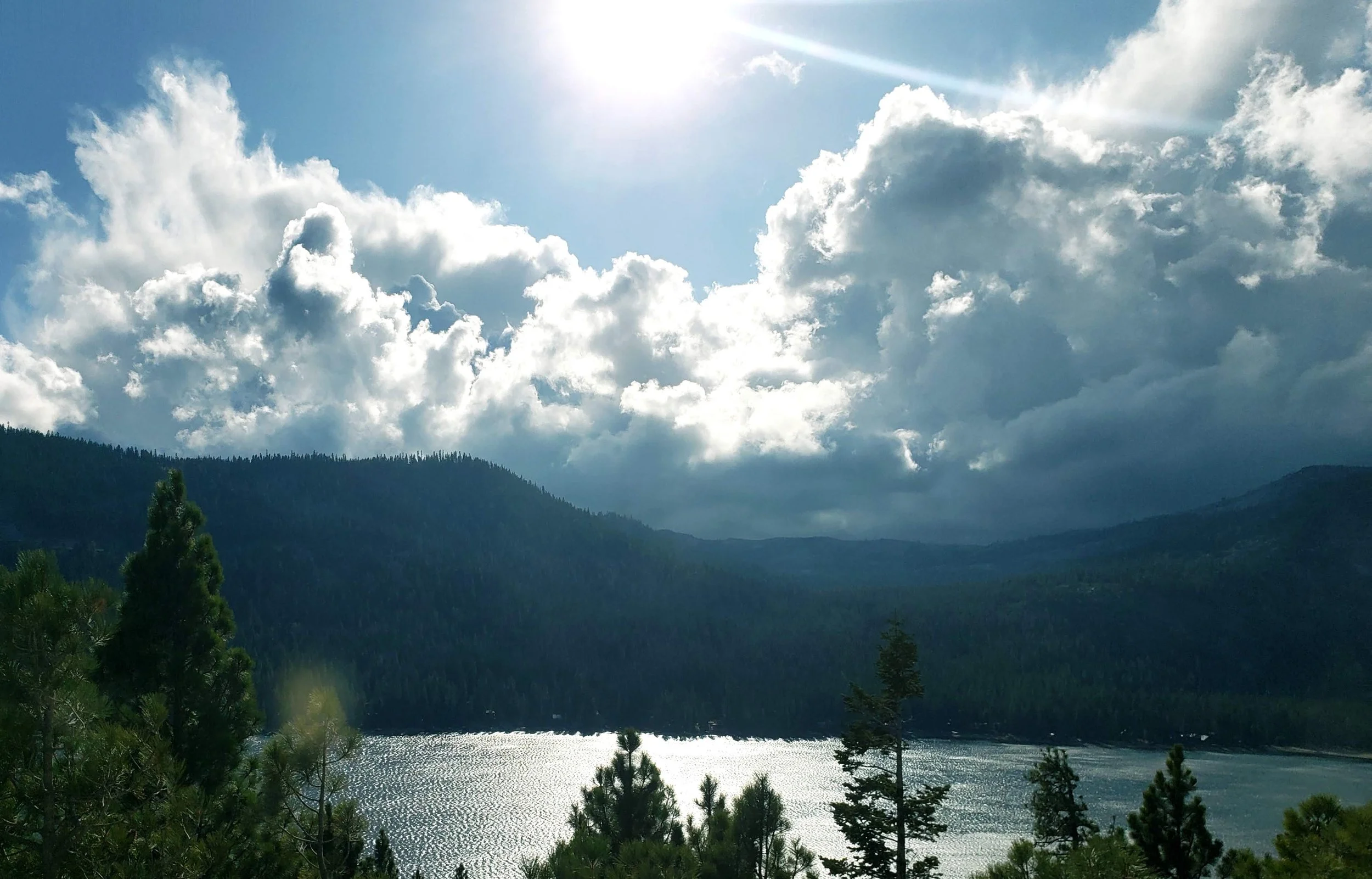 A scenic view of a lake surrounded by forested mountains, with a partly cloudy sky and sunlight shining through clouds.