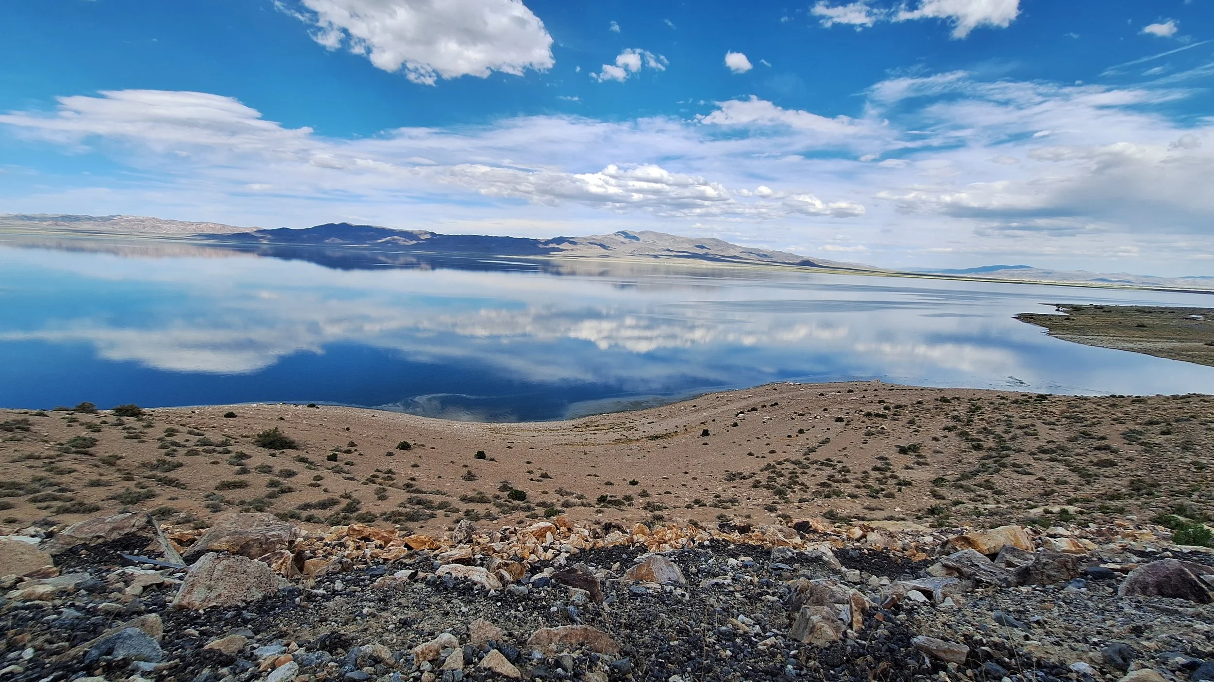 A calm lake with mirror-like water reflects the sky and clouds above. The shoreline is rocky and arid with sparse bushes, and distant mountains are visible under a partly cloudy sky.