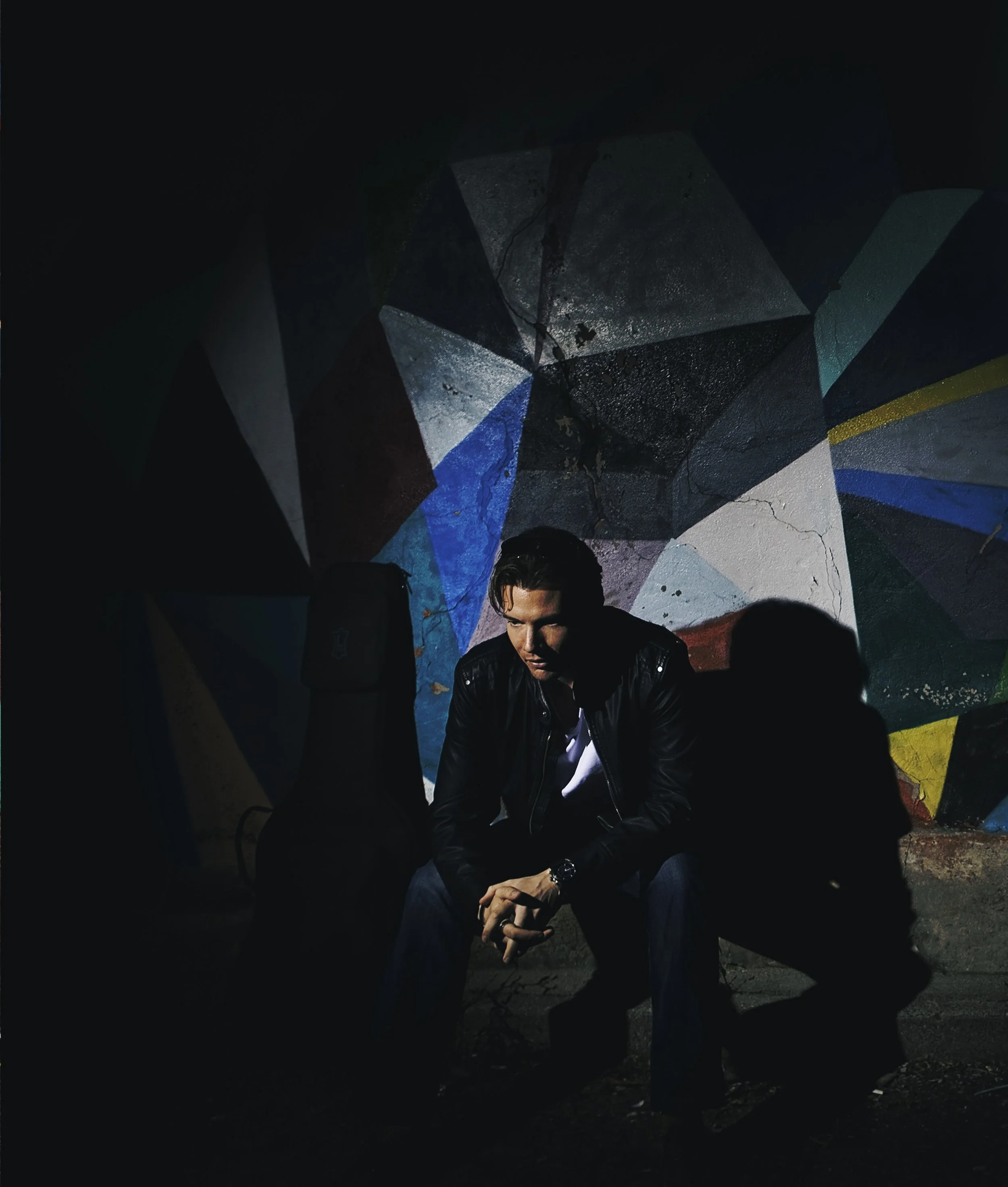Sean Hayden sitting on a bench in front of a colorful geometric mural at night, wearing a black leather jacket and a watch, with a guitar case beside him.