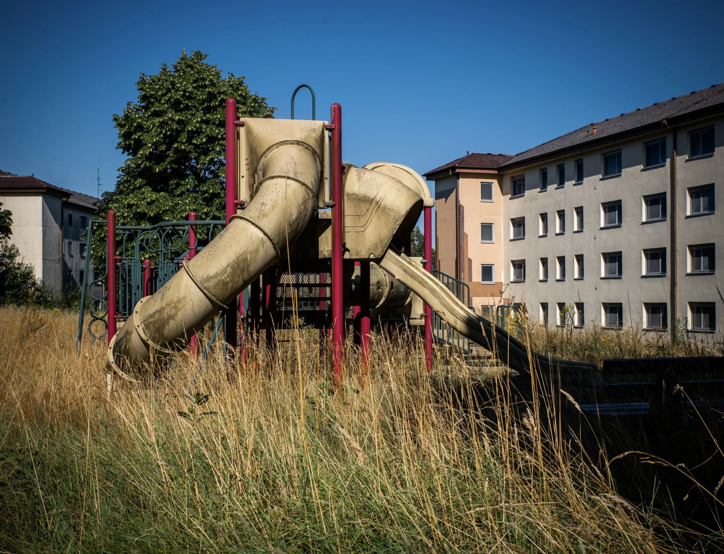 Verwachsener Spielplatz mit verrosteter Rutsche und Klettergerüst, umgeben von hohem Gras, vor Wohngebäuden unter blauem Himmel.