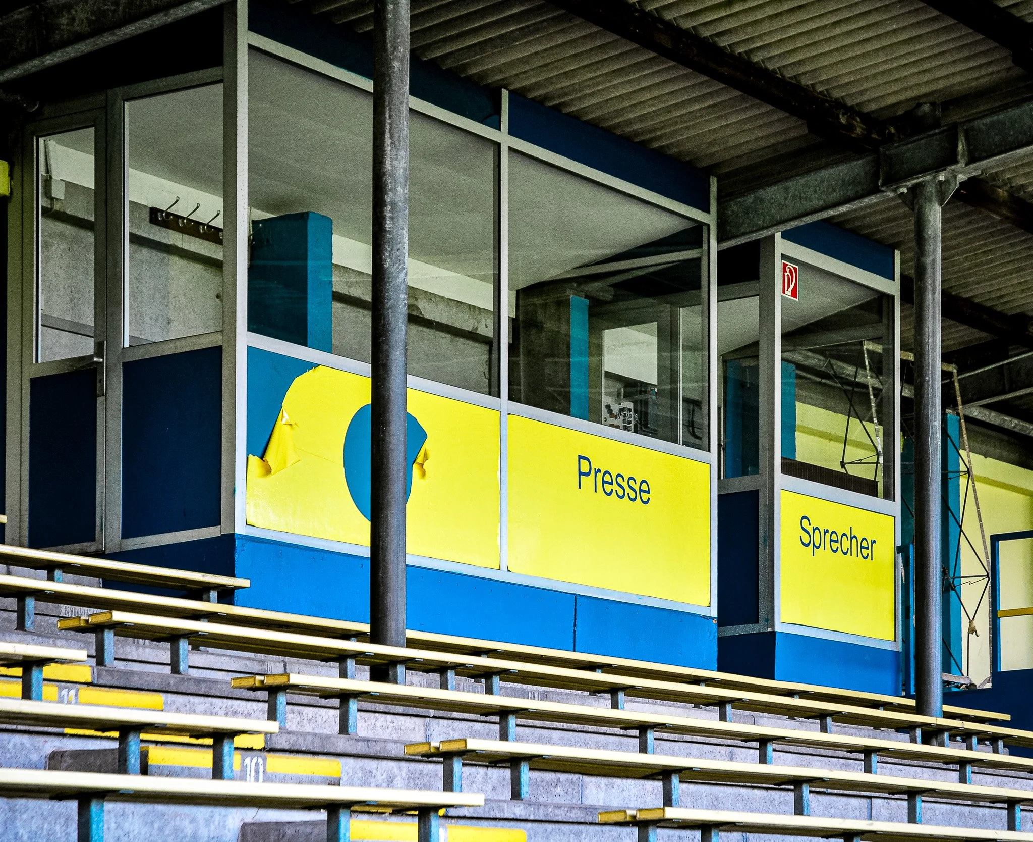 Eine leere Stadiontribüne mit gelben und blauen Elementen. Im Hintergrund sind Kabinen mit der Beschriftung "Presse" und "Sprecher" zu sehen.