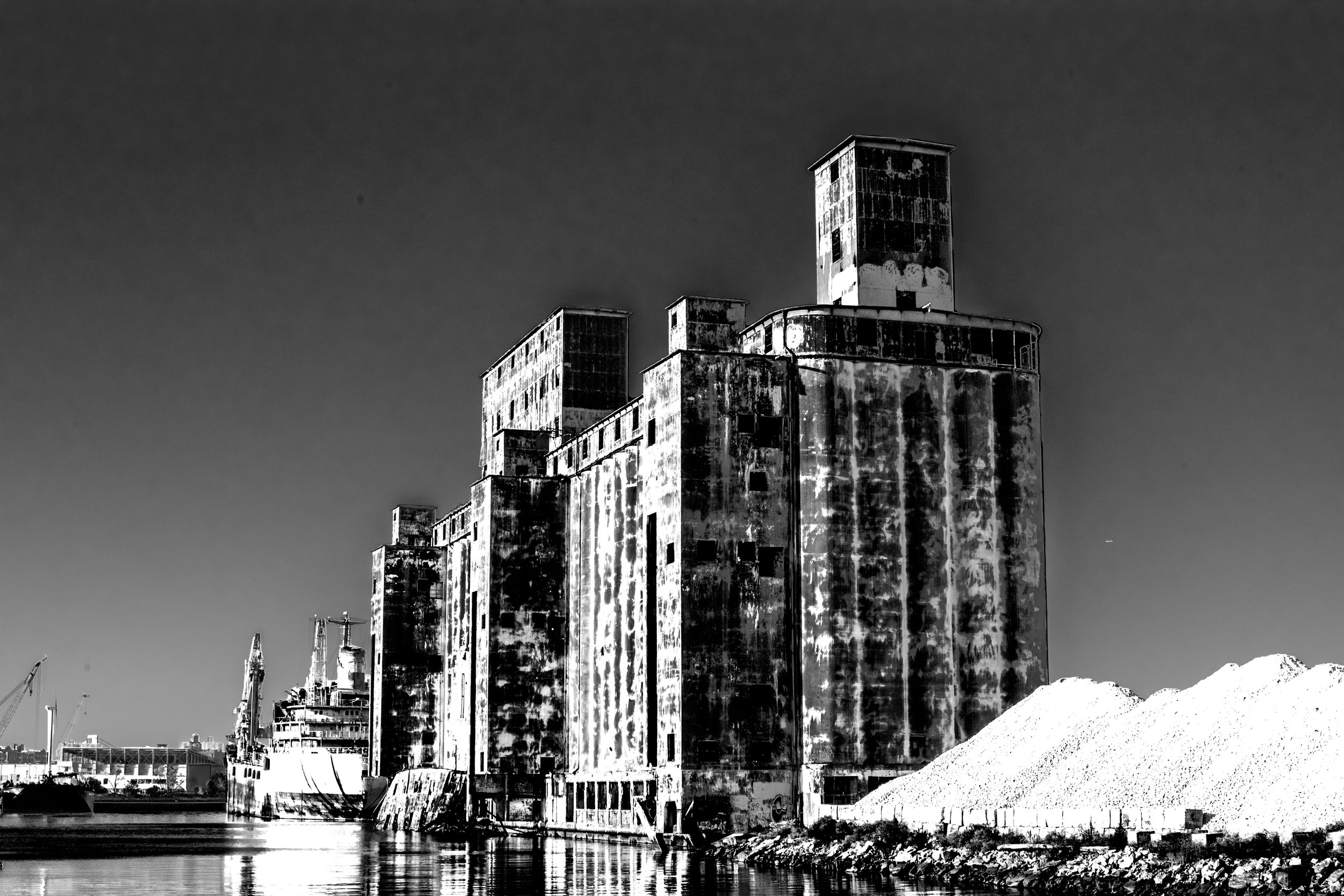Black and white photo of tall, old, weathered industrial buildings along a waterfront with boats docked nearby.