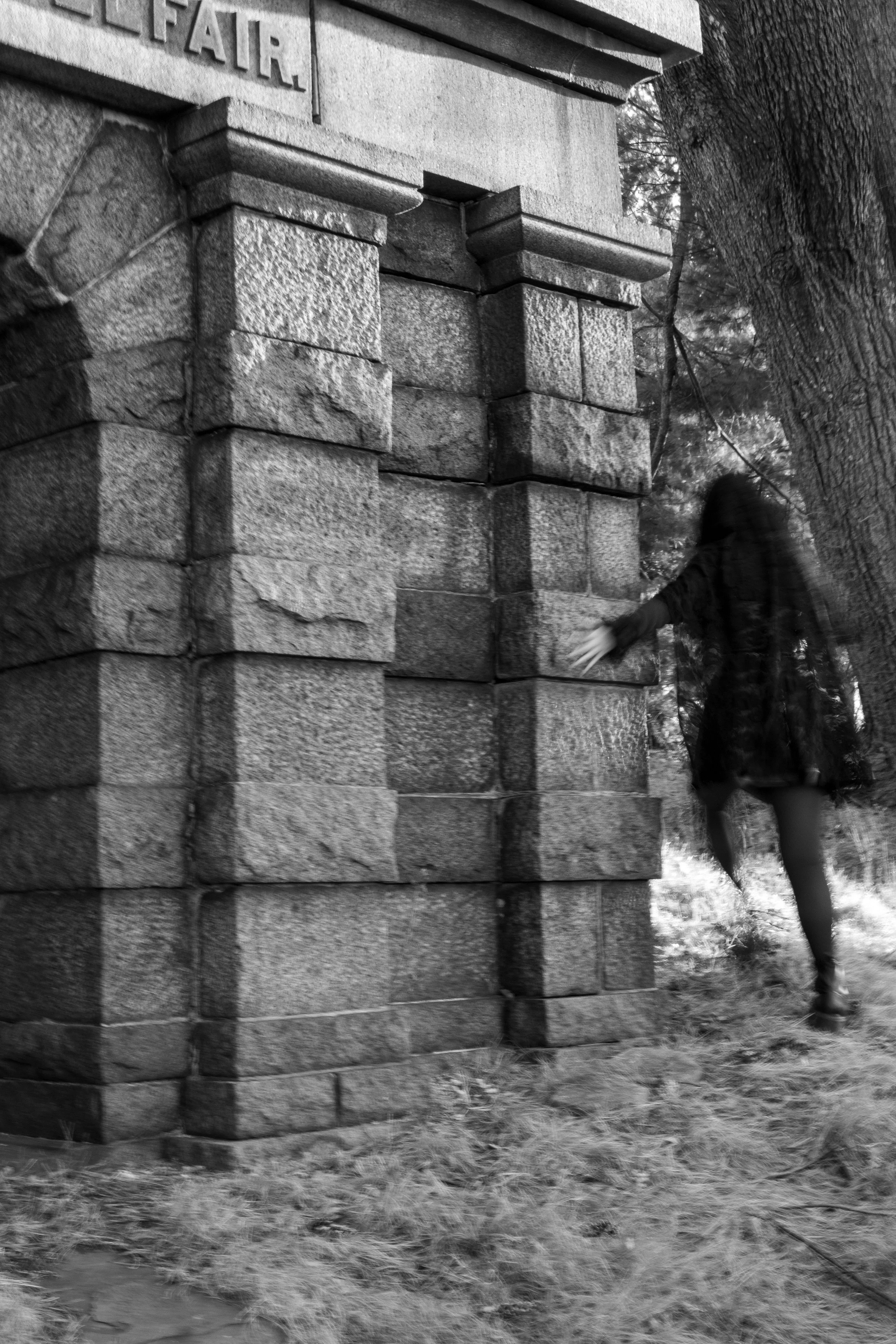 Black and white photo of a stone monument with the word 'FAIR' partially visible on it, trees in the background, and a person walking by.