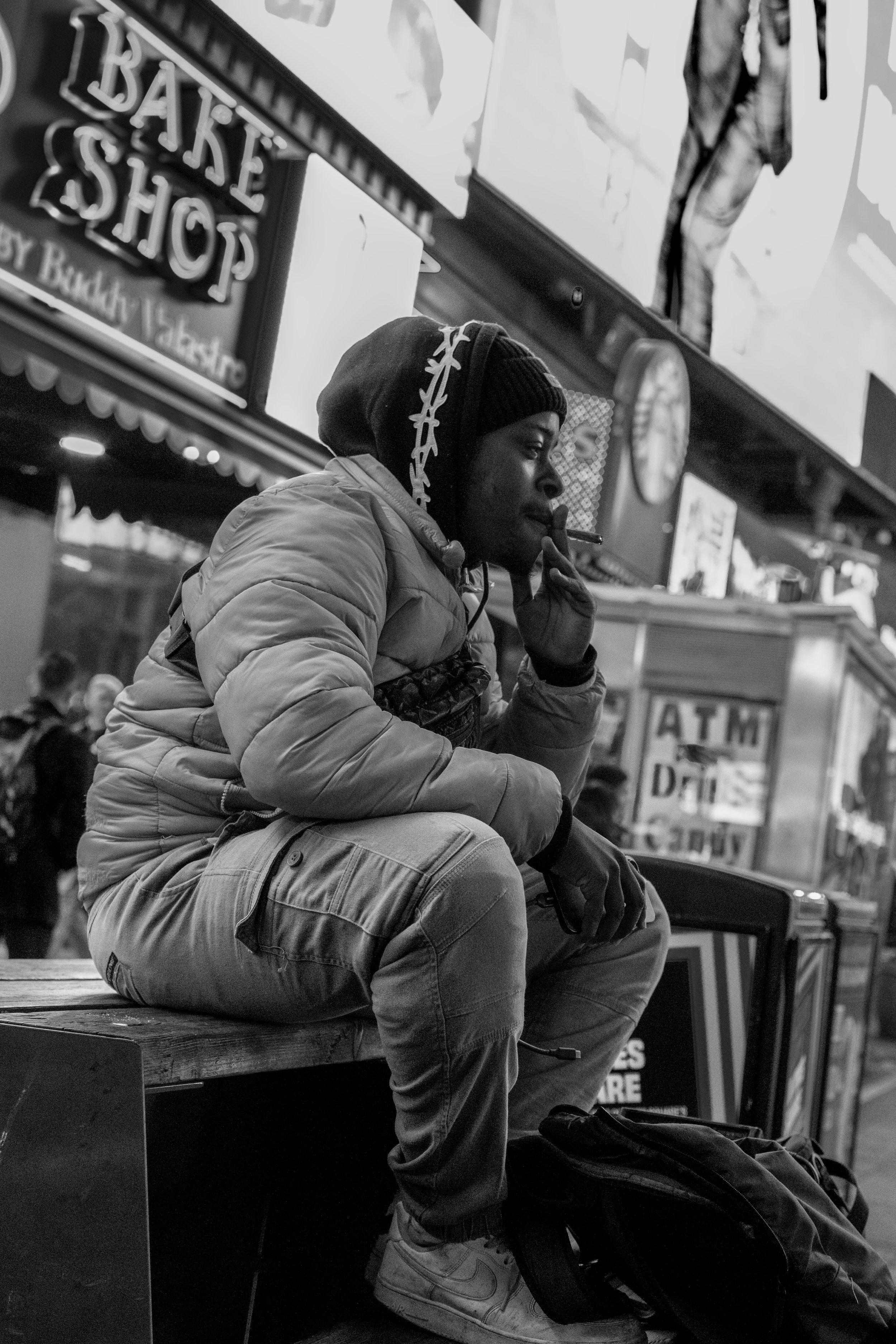 A young man sitting on a bench in front of a shop, smoking a cigarette. He's wearing a winter jacket, a beanie, and sneakers.
