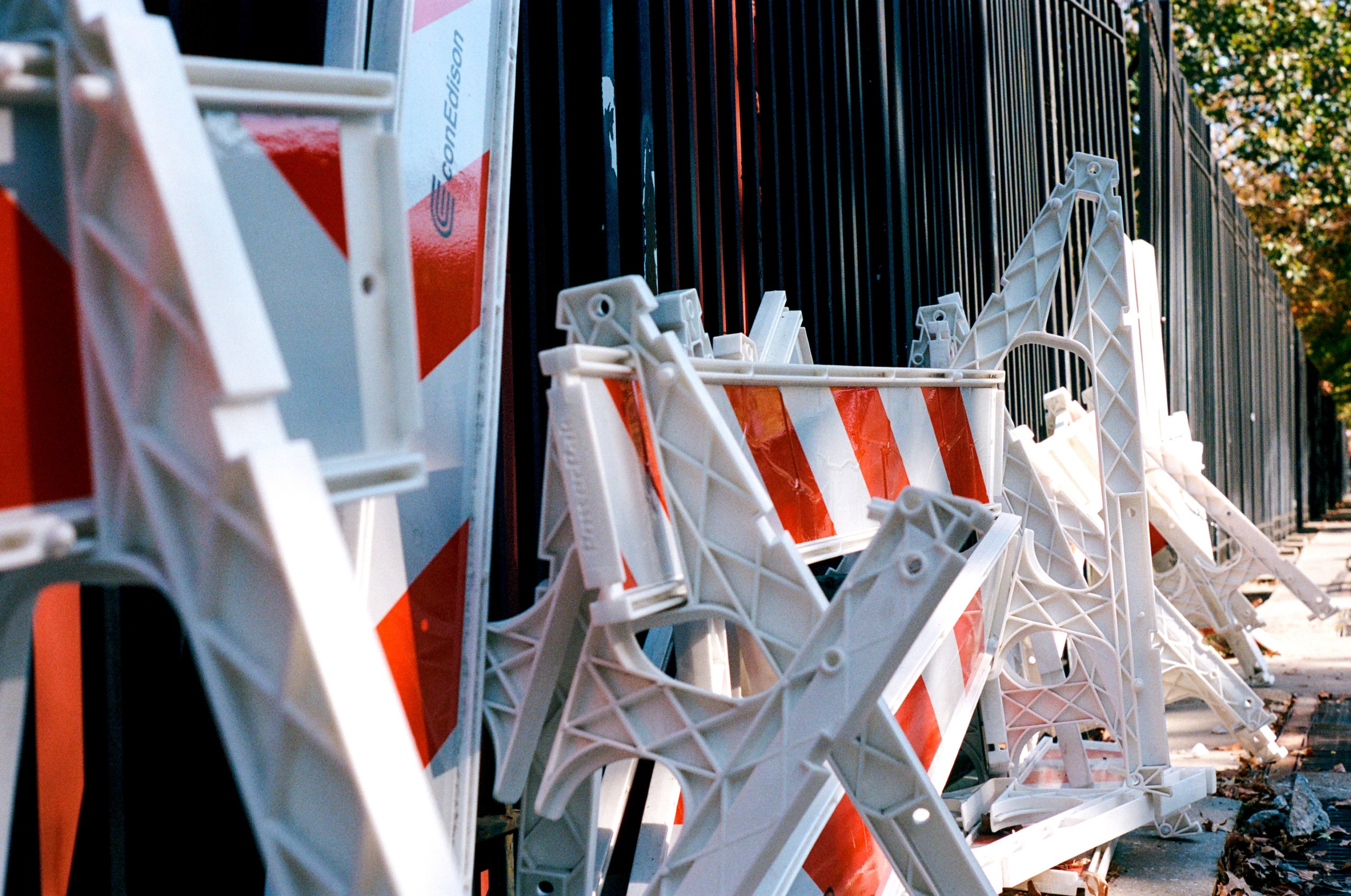Row of orange and white striped barricades leaning against a black fence with trees in the background.