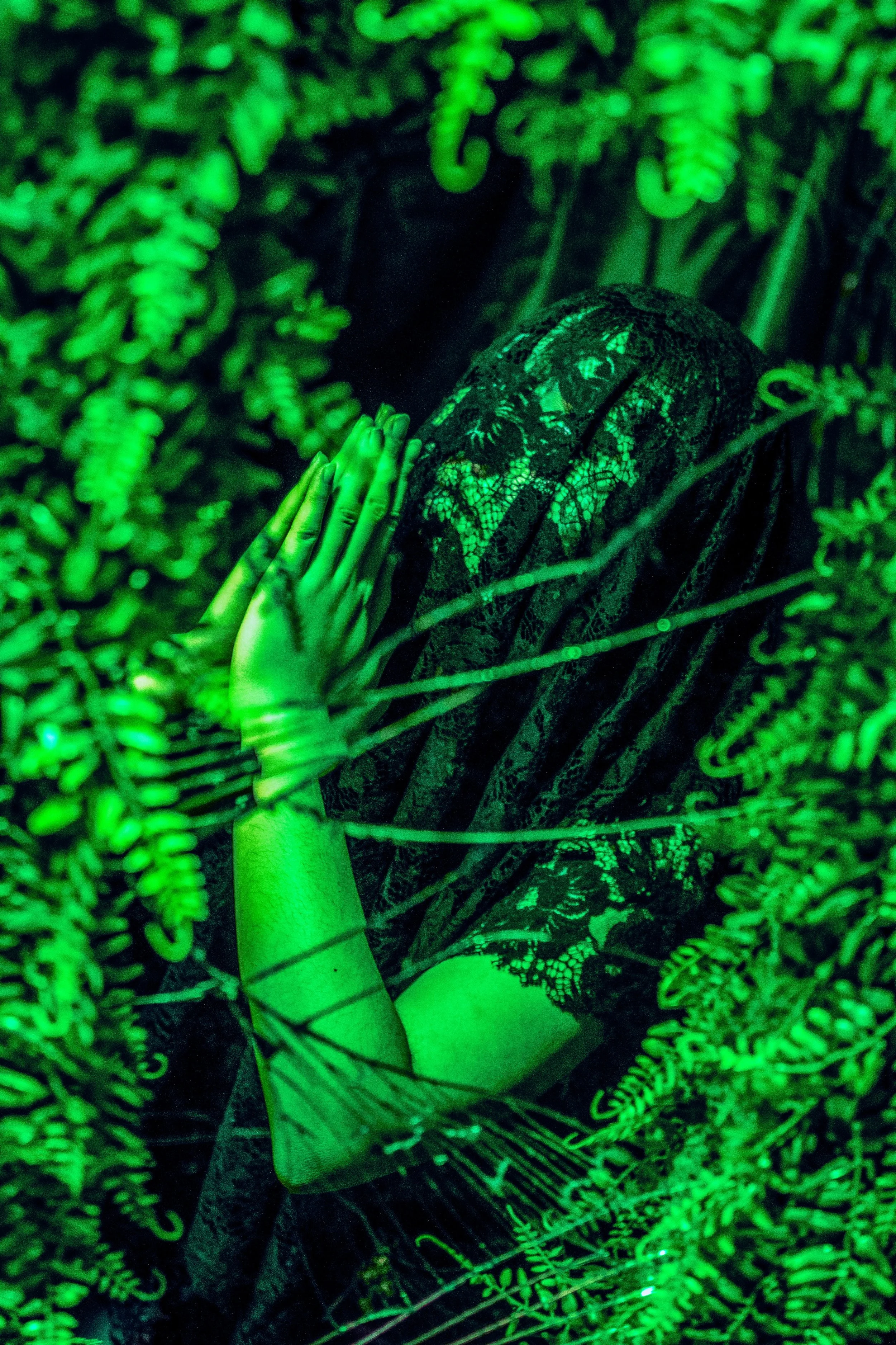 A person is in a dark forest surrounded by green glowing plants and ferns. They are wearing a patterned outfit, with one hand pressed against a tree or branch and the other hand resting on their face, looking upward.
