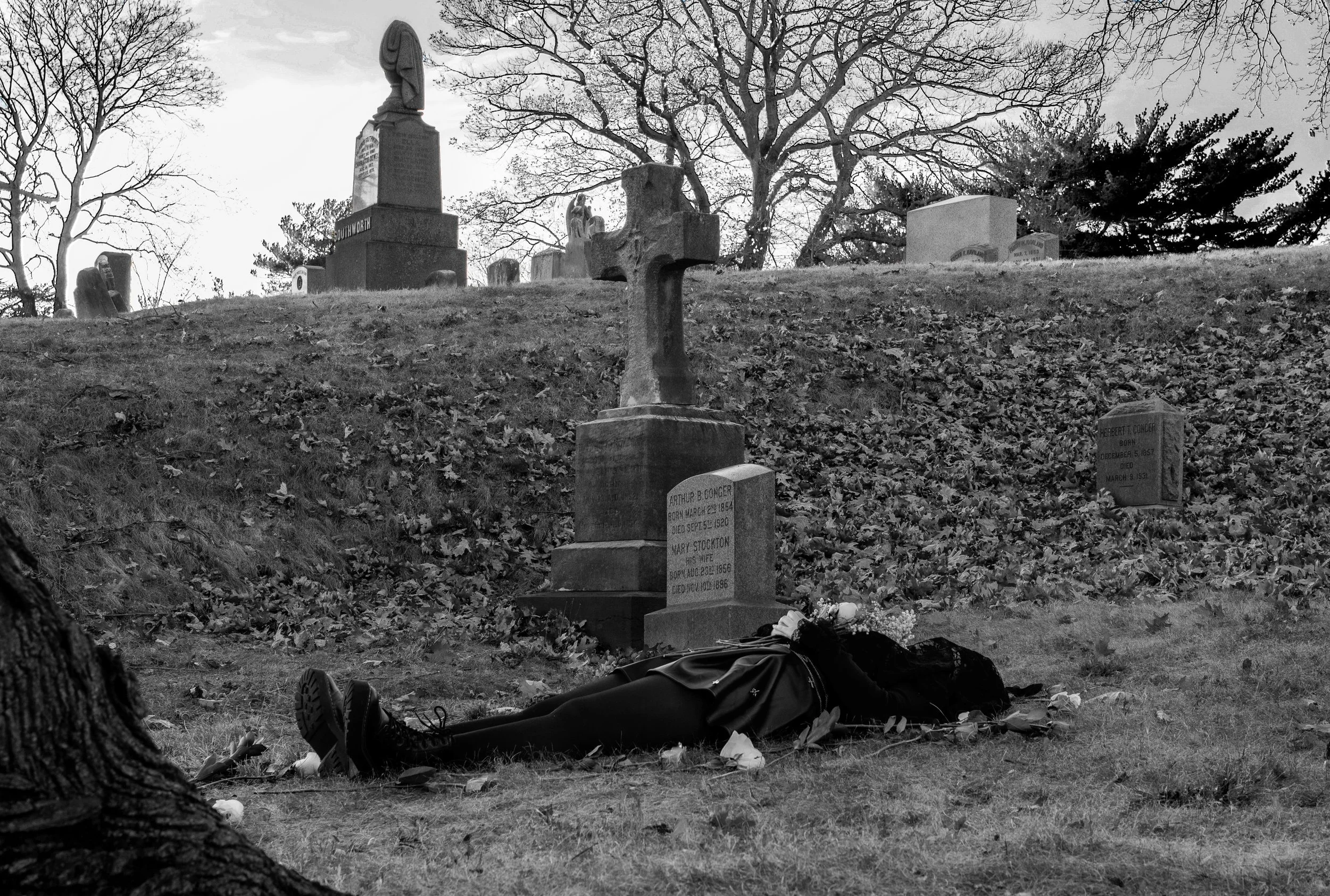 A person lying on the ground in a graveyard, wearing dark clothing and boots, with their head towards a tombstone. Several tombstones are visible, and leafless trees are in the background on a cloudy day.