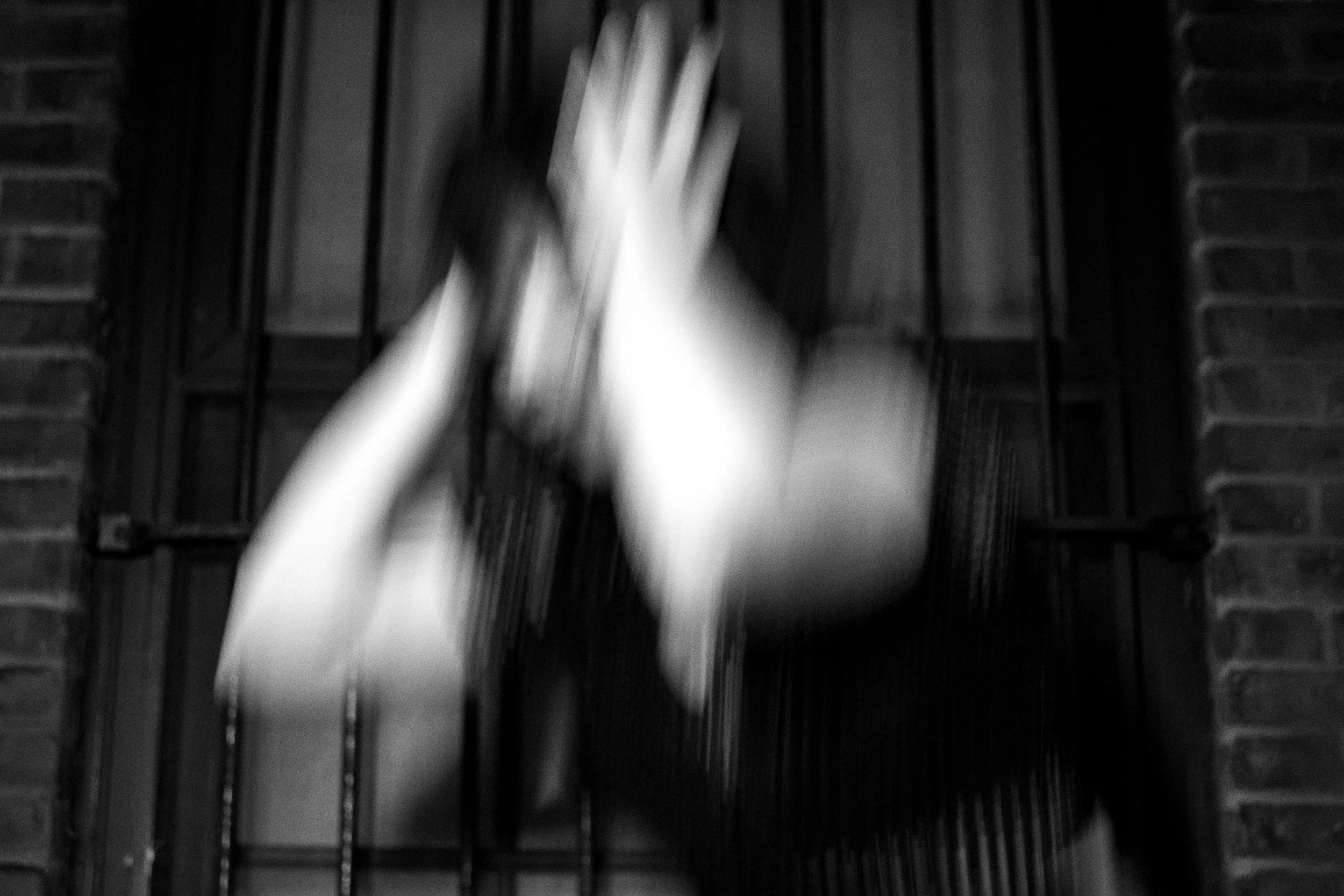 Black and white photo of a person reaching up with both hands, standing in front of a brick wall with a window and black metal bars.